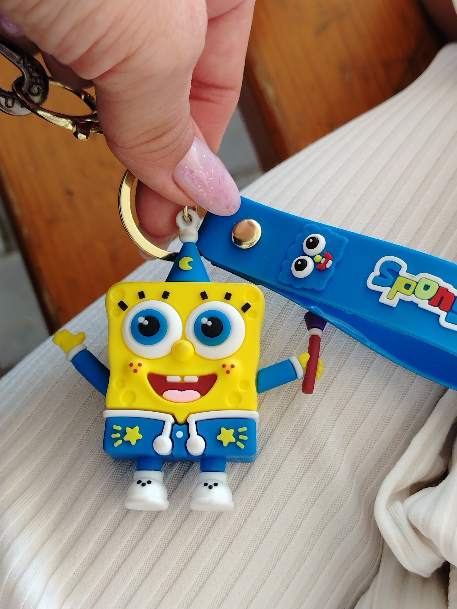 A hand with pink glitter nail polish is holding a keychain. The keychain features a yellow, rectangular, 3D cartoon figure with a blue hat. The figure has two large white eyes with black pupils, a red mouth, yellow arms and legs, and a blue outfit. It appears to be SpongeBob SquarePants in a modified design. A blue rubber strap attached to the keychain reads "Sponge" in white lettering with green and red accents, and a blue monster-like face. The background includes a wooden surface and a white textured fabric. The location is not identified.