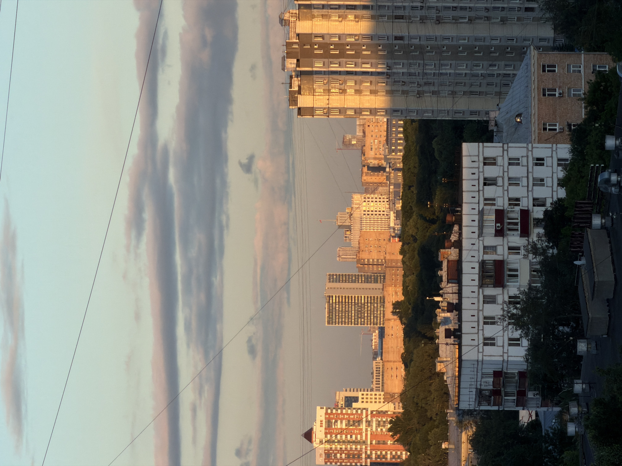 The image shows a cityscape in Khimki, Russia, during what appears to be either sunrise or sunset, given the warm golden light illuminating some of the buildings. In the foreground, a multi-story apartment building with white siding and dark red balconies is visible. To its right, a taller, grey apartment building with numerous windows dominates the frame, reflecting the golden light. Further in the background, a cluster of various buildings of different heights and architectural styles can be seen, interspersed with trees and greenery. Power lines stretch across the sky, which is a soft gradient of blue and pink from the clouds. There are no people or visible actions occurring in the scene. The overall impression is a residential urban environment bathed in the soft glow of the sky.