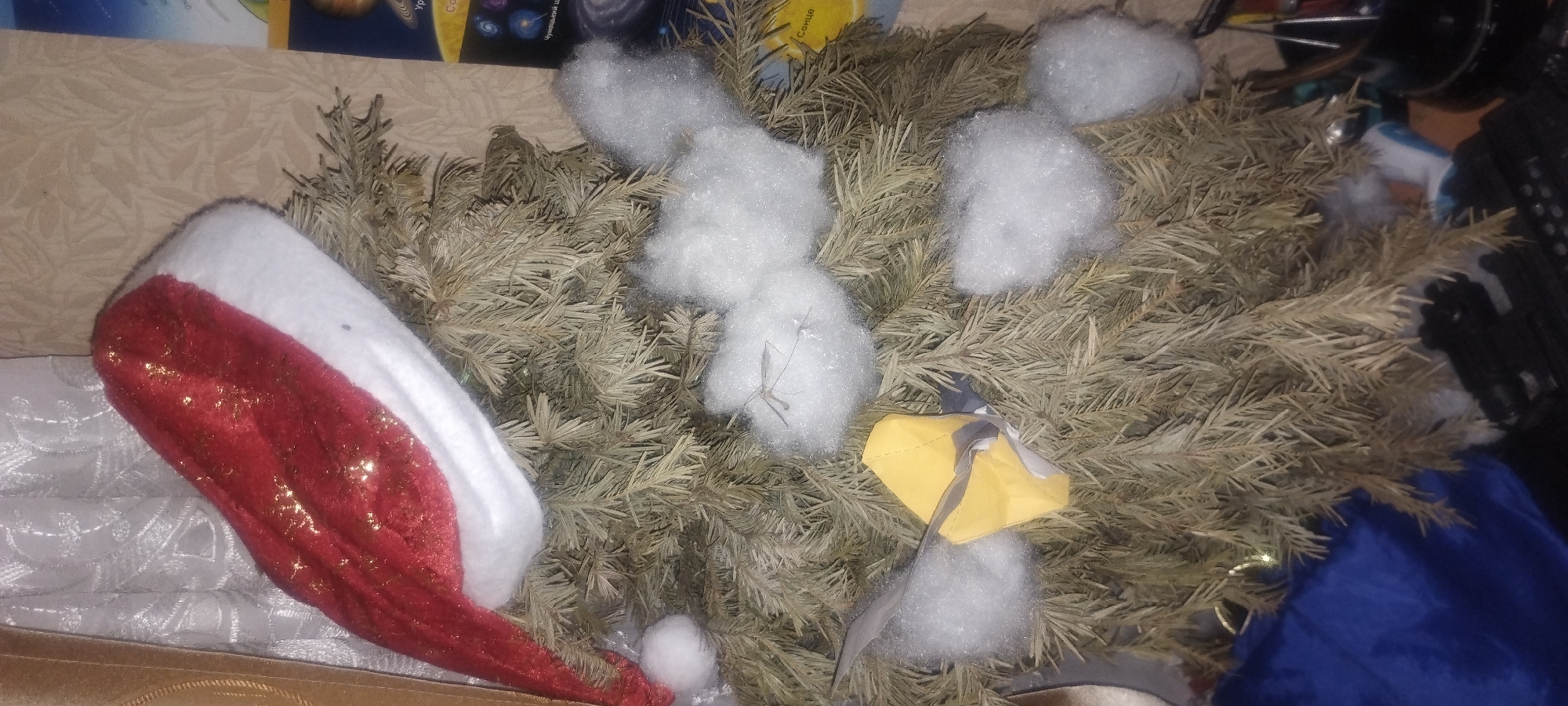 The image shows a close-up of a small, artificial Christmas tree adorned with a Santa hat and some artificial snow. A small yellow object, possibly a gift tag or ornament, is attached to the tree. The tree is decorated with what appears to be cotton balls as snow. In the background, a wall with a patterned wallpaper is visible, along with a glimpse of what looks like an educational poster about space. A blue fabric is present at the bottom left of the frame. The scene appears to be indoors, likely in a residential setting. There is no identifiable information or context to determine its location within the city of Zaporizhzhia, Ukraine.