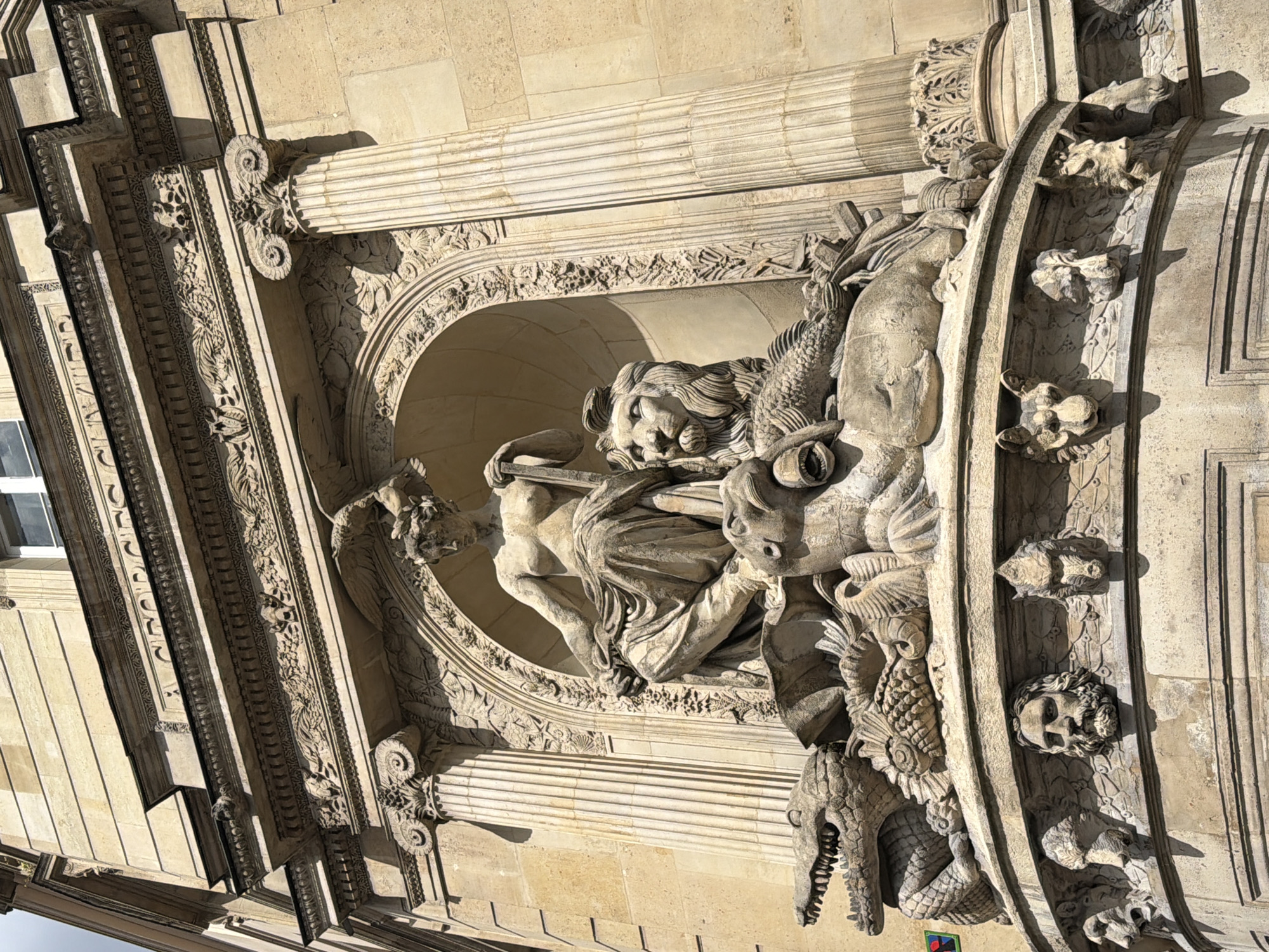 A stone-carved sculpture adorns the facade of a grand building, likely in Paris, France, captured on a bright, sunny day with strong shadows. The image is rotated 90 degrees clockwise.
The central feature is a classical-style female figure, possibly an allegorical representation or a goddess, within an arched niche. She is depicted with flowing drapery, kneeling or seated, and appears to be holding a long, slender object, possibly a spear or staff, pointing upwards and to the left (when viewed correctly). Her gaze is directed upwards and to the right.
Surrounding the central figure are numerous ornate stone carvings. To her right (our left in the rotated image), a lion's head with a flowing mane is prominent. Below and around her are various marine and fantastical creatures, including fish, dolphin-like forms, and grotesque heads of dragons or other beasts with sharp teeth, integrated into the arch's spandrels and columns.
The sculpture is set within an elaborate architectural framework featuring fluted columns with Ionic capitals, richly decorated friezes with intricate floral and shell patterns, and moldings. The building itself is constructed from light-colored, cut stone blocks. Numerous smaller decorative heads and figures are carved into the curved elements flanking the central arch.
There is no discernible legible text visible in the image.