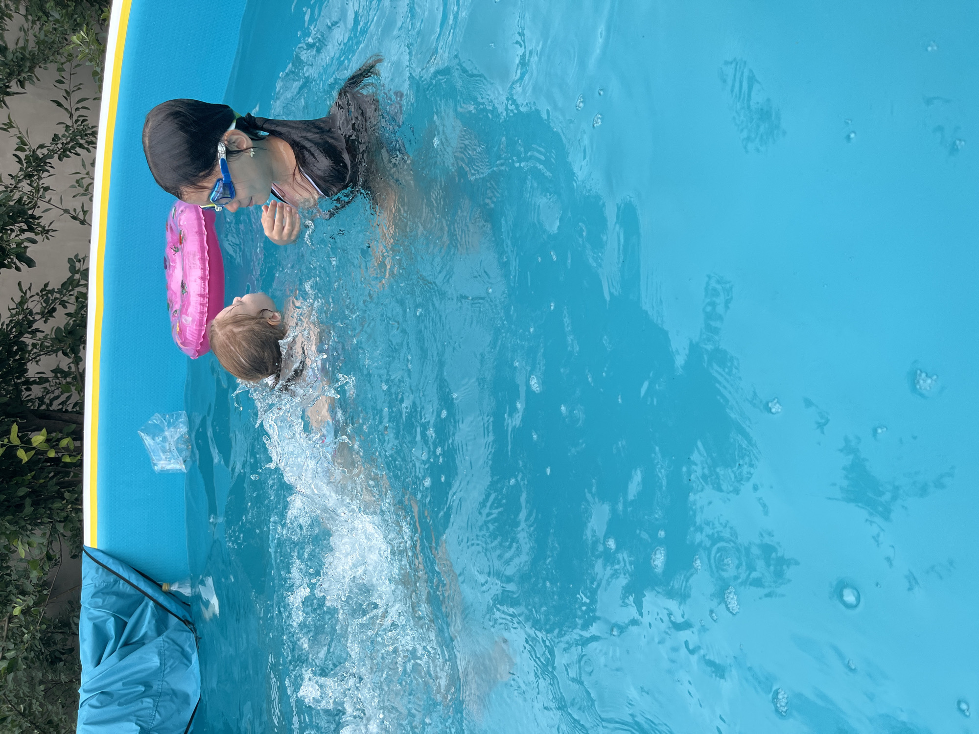 In a bright blue, inflatable swimming pool, two young girls are engaged in playful activity. The girl on the left, with blonde hair and a fair complexion, is actively splashing water, her body partially submerged and creating a significant water disturbance. She appears to be swimming or moving vigorously through the water. The girl on the right, with dark hair pulled back into a ponytail, is wearing blue swimming goggles and a dark colored swim shirt. She is looking towards the other girl, seemingly interacting with her. A pink inflatable ring with a donut-like pattern floats behind the blonde girl, suggesting a recreational atmosphere. The pool is situated outdoors, as indicated by the presence of green foliage and trees in the background. The scene captures a moment of summer fun and sibling interaction within a residential or recreational setting. There is no specific identifiable information to place this image within Shymkent, Kazakhstan.