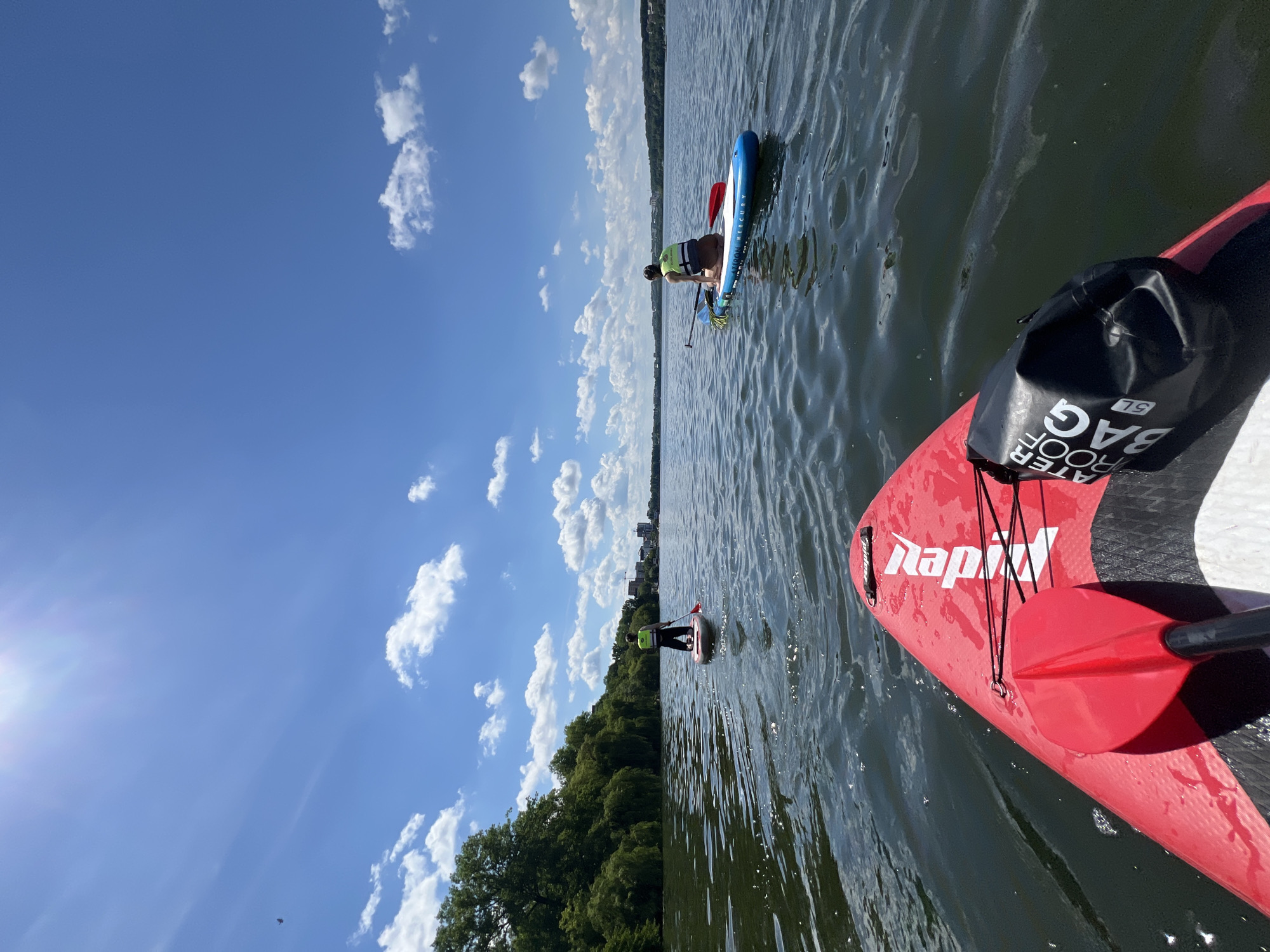Two people are stand-up paddleboarding on a calm lake under a bright blue sky with scattered white clouds. The perspective is from the front of a red paddleboard in the foreground, looking out towards the other paddleboarders. The person in the foreground is facing away from the camera, kneeling on their board. They are wearing a light green life vest and brown shorts. Another person is further in the distance, standing upright on a blue and white paddleboard. They are also wearing a light green life vest. Both individuals are holding paddles. The background features a tree-lined shore with buildings visible in the distance. The water is rippling, reflecting the sky. The scene appears to be the Ternopil Pond in Ternopil, Ukraine, given the characteristic shoreline and city skyline.