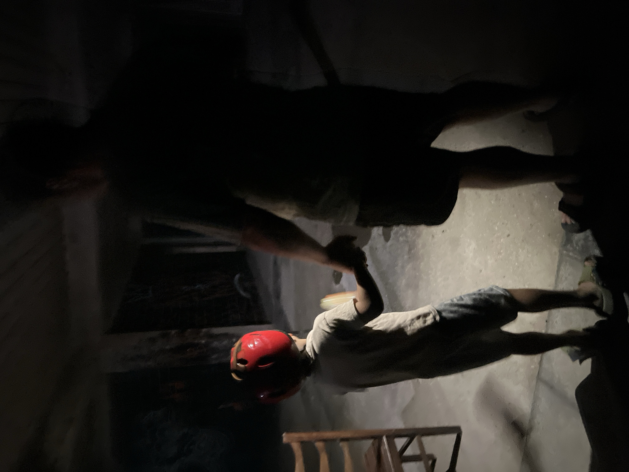 In Poti, Georgia, in what appears to be a dimly lit outdoor area with a concrete floor and overhead structure, a person, likely an adult, holds the hand of a child. The child is wearing a red helmet and a light-colored t-shirt, and appears to be holding a small, flat object in their other hand. To the left of the child, a wooden chair is visible. The lighting suggests it is either dusk or nighttime.