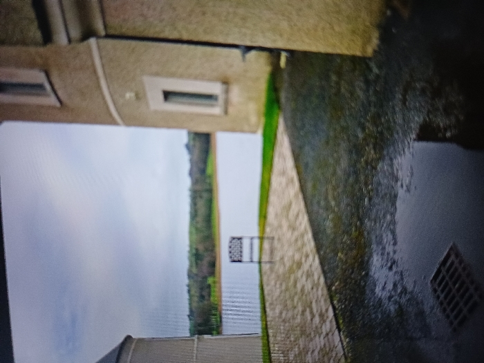 The image depicts an outdoor scene at the Lough Erne Resort in Enniskillen, County Fermanagh, United Kingdom, overlooking Lough Erne under an overcast sky. In the foreground, a dark, wet gravel or loose stone surface with a prominent puddle occupies the right and lower left. A square metal grate is visible within the puddle in the bottom left corner. To the left of the wet area, a paved patio section with light-colored, rectangular pavers laid in a herringbone or similar pattern is present. A single dark-colored, possibly metal, outdoor chair with an open-patterned backrest stands centrally on this paved area, facing the water.
To the left, part of a light-colored, rounded building or tower structure is visible. To the right, a substantial building with light-colored rendered walls features a rounded section with two narrow, vertical, recessed windows with light frames. A dark vertical drainpipe runs down the right side of this building. Between the paved area and the building on the right, a strip of green grass is visible.
In the mid-ground, a wide expanse of calm water, identified as Lough Erne, stretches horizontally. Beyond the lough, a distant treeline and undulating green landscape are visible under a uniformly cloudy, bright grey sky. No individuals or active subjects are present. The scene conveys a quiet, damp, and unoccupied waterside environment.