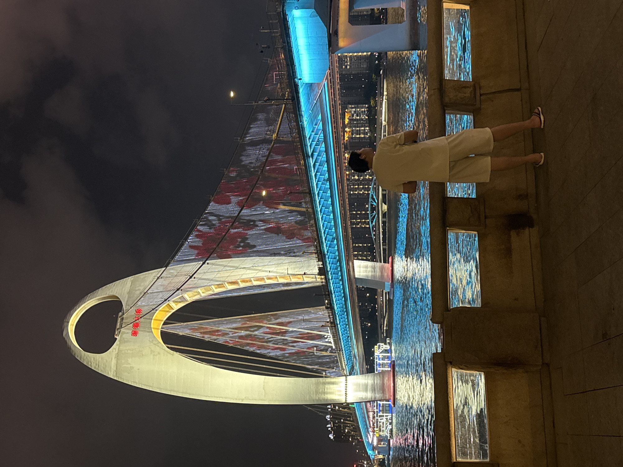 A nighttime shot in Tianhe District, China, showcases a large, illuminated bridge over water. The bridge features a distinctive arch structure and a decorated roadway, likely with LED lights. A person in light-colored clothing stands on the walkway beside the water, facing the bridge, but their activity is indiscernible. The surroundings include buildings and urban lighting visible across the water, with an overcast sky forming the backdrop.