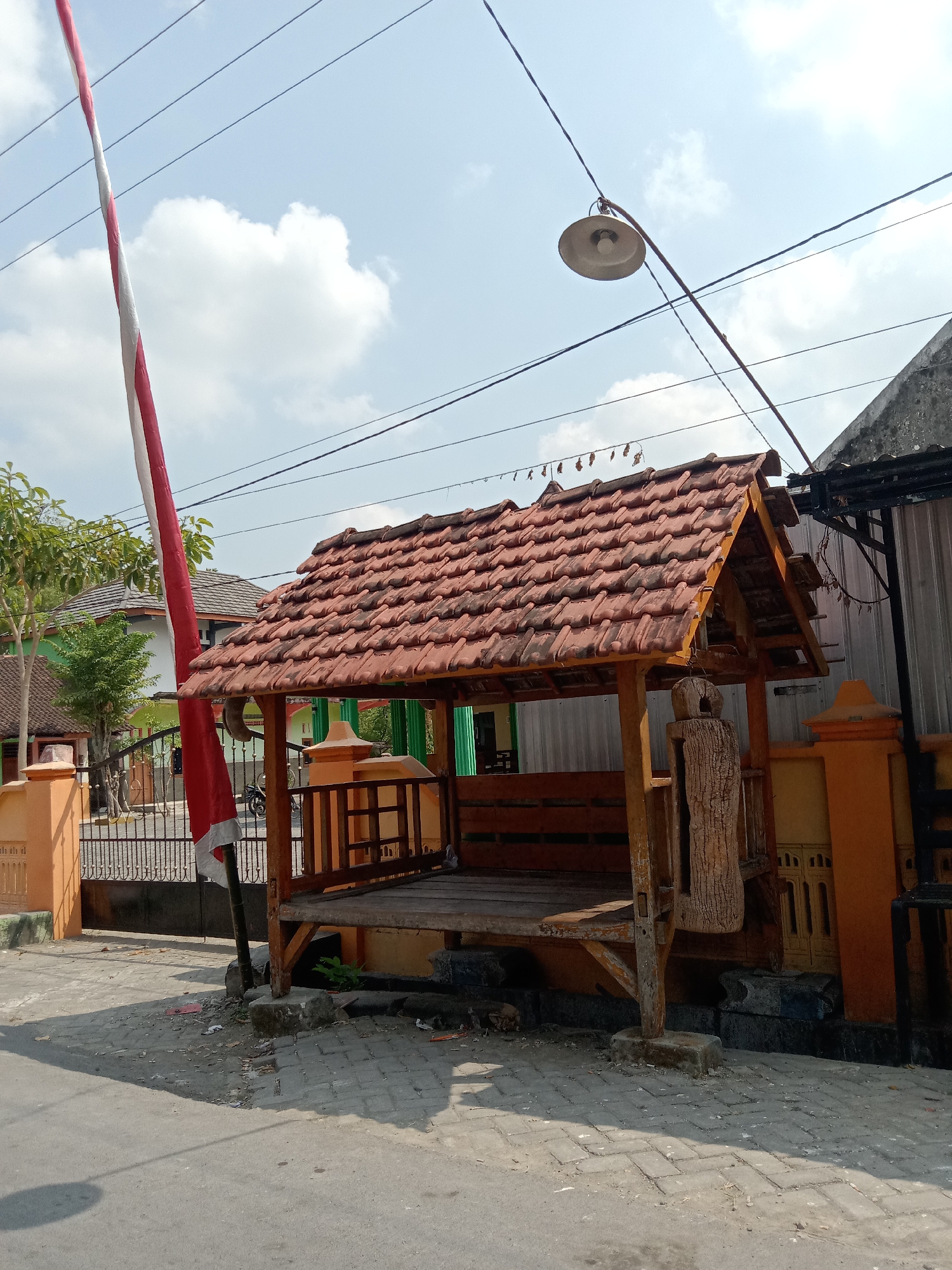 A wide, low-angle, outdoor shot shows a traditional Indonesian gazebo with a red-tiled roof. The gazebo is made of wood and has benches inside. A red and white Indonesian flag is visible on the left side of the frame, attached to a pole. In the background, there are houses with similar tiled roofs and some trees. Overhead, power lines and a street lamp are visible against a partly cloudy blue sky. The ground in front of the gazebo is paved with grey bricks. The scene is likely located in a residential area in Sugihwaras, Indonesia.