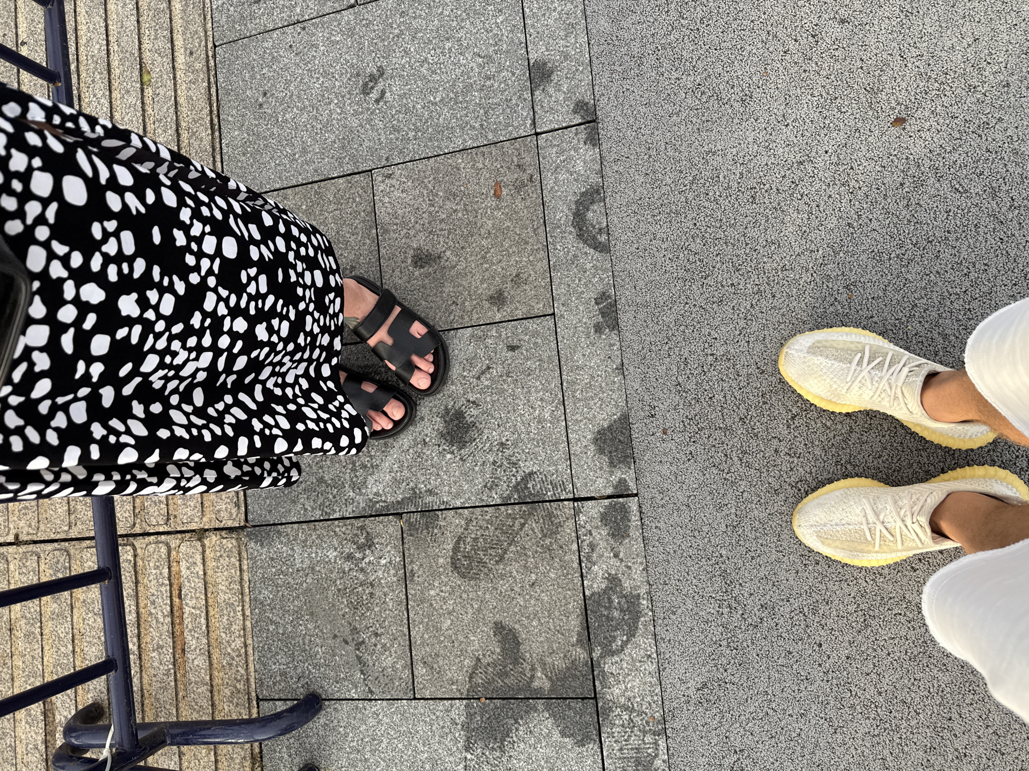 Two people stand on a paved walkway in Haizhu District, China. The person on the right wears a black and white spotted dress and black sandals. The person on the left wears white shorts and cream-colored sneakers. The ground is made of gray stone tiles and a rougher gray aggregate material. There are water stains on the stone tiles. A blue metal railing is visible in the upper left corner. The lighting suggests it is daytime, possibly late afternoon due to the presence of shadows.