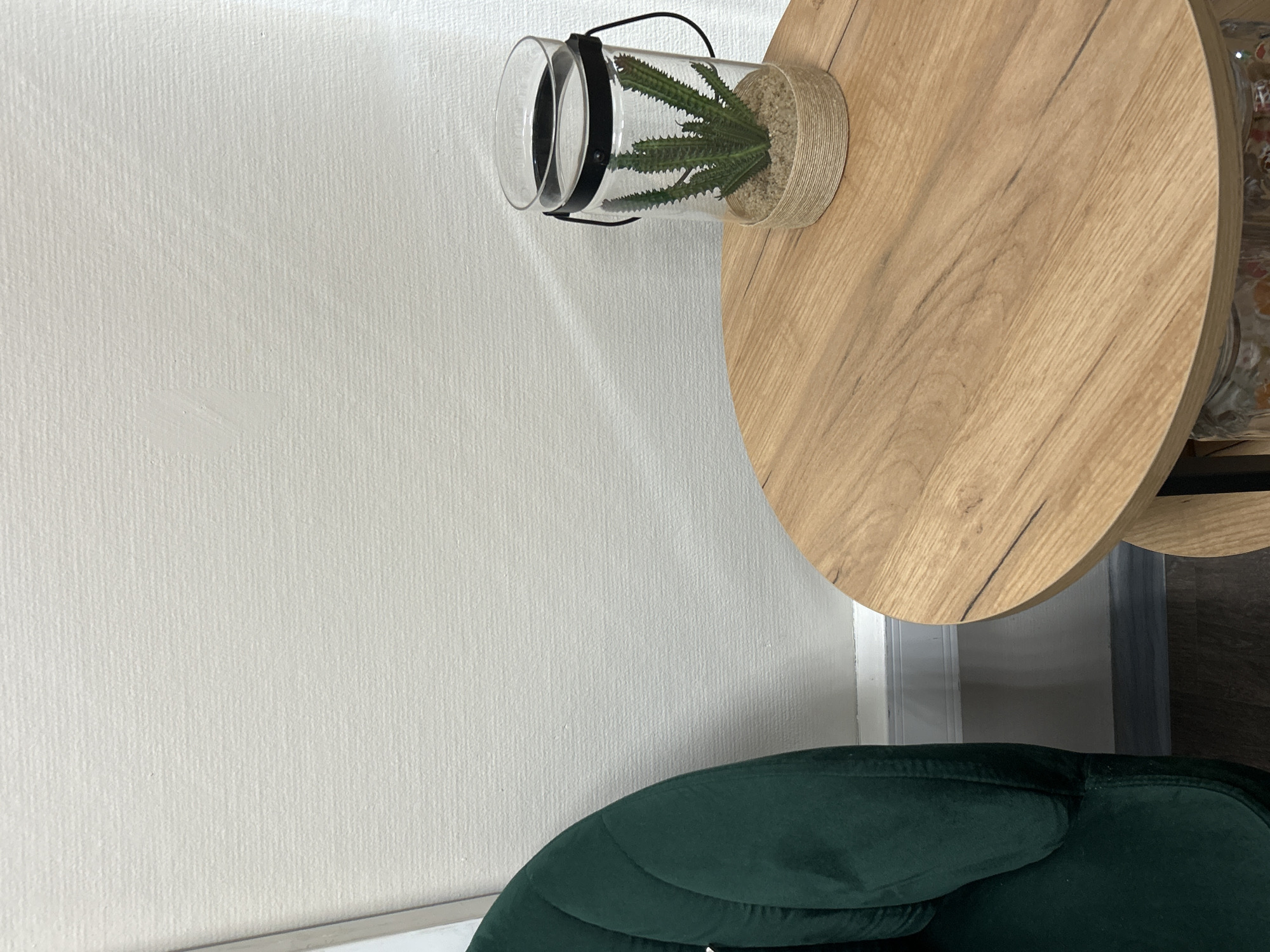 The image depicts an indoor scene with a partial view of a dark green upholstered chair, a light wood round side table, and a decorative plant. On the left, a segment of a dark green, possibly velvet or plush, chair with a visible seam or channel is present. To its right, a round wooden side table with a light brown wood grain finish featuring darker streaks and knots is centered. On the table, a clear cylindrical glass container holds a faux green cactus plant embedded in light brown sand or pebbles. The top rim of the glass container is encircled by a black metal band with a thin black loop handle, and the bottom exterior is wrapped with natural jute twine. The background consists of an off-white or light beige wall with a subtle horizontal textured pattern. A white baseboard or molding runs along the bottom of the wall, above a dark, possibly wood laminate, floor. No individuals are present, and no actions are occurring. The location is an interior space in the city of Unknown, Unknown.