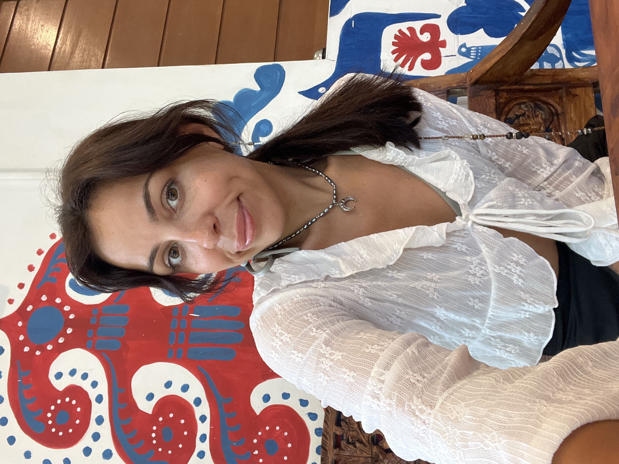 A woman with long, dark brown hair and warm brown eyes looks directly at the viewer with a subtle, playful smile, her tongue just peeking between her lips. She is wearing a white, semi-sheer, long-sleeved top adorned with delicate lace patterns and ruffles, tied loosely at the front. Around her neck, she wears two necklaces: a shorter one with dark beads and a silver crescent moon pendant, and a longer one crafted from a brown cord featuring various small, multi-colored beads.
The background is a vibrantly painted wall or panel featuring a folk-art style design in bold red, blue, and white. The intricate patterns include large swirling motifs, dots, and stylized figures, possibly resembling birds or abstract shapes. Part of an ornately carved wooden chair or furniture is visible in the upper right quadrant of the image.
The lighting is bright, suggesting a daytime setting, possibly indoors or in a well-lit shaded area. The overall impression is one of warmth and cheerfulness, conveyed by the woman's engaging expression and the colorful surroundings. The artistic background and carved wood hint at a culturally rich environment, aligning with the location context of Kuta Selatan, Indonesia. There is no visible text in the image.