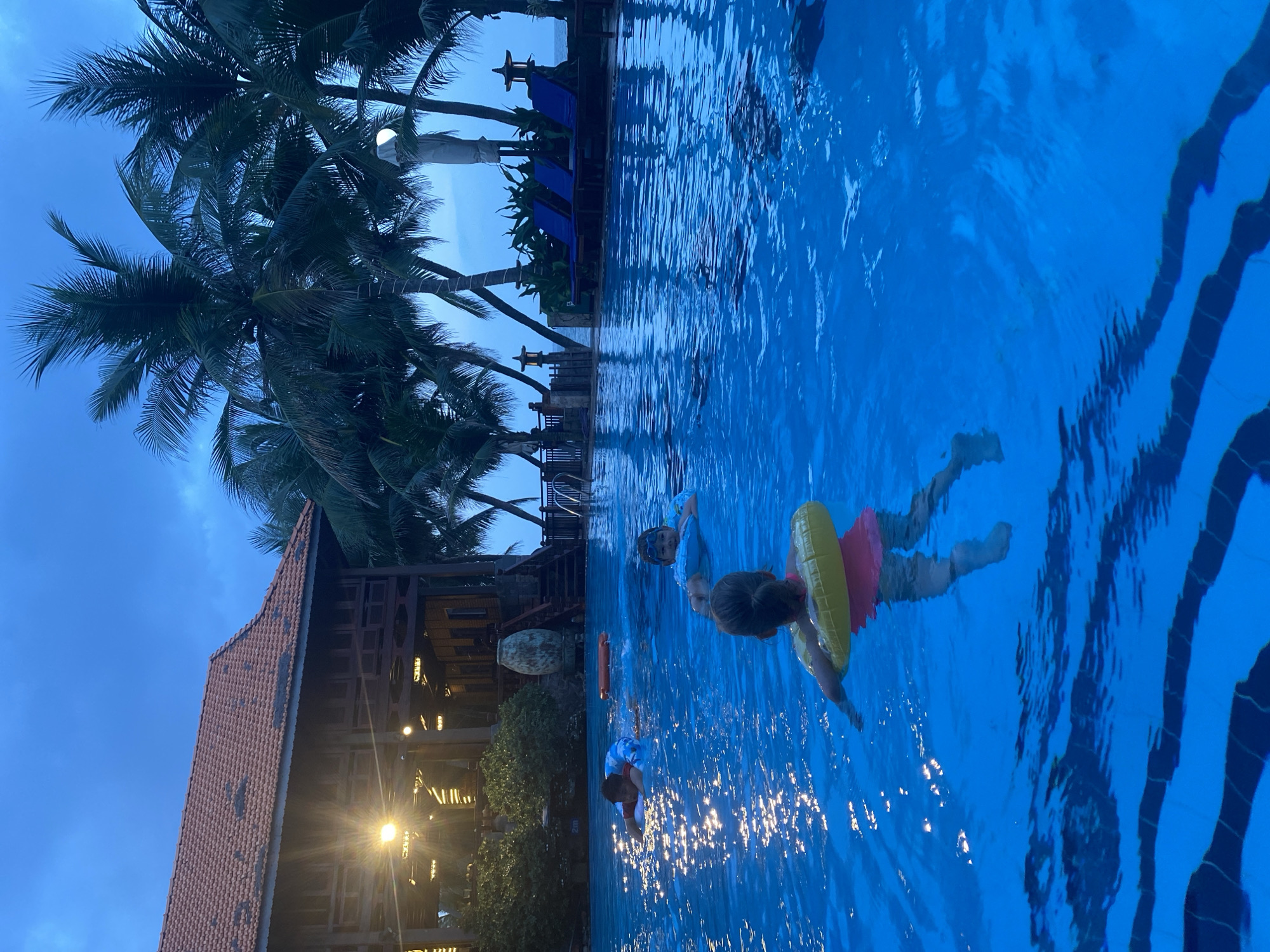 This image depicts an outdoor swimming pool at dusk, with a multi-story wooden building with a red tiled roof in the background. Several palm trees are visible, and the ocean can be seen in the distance. Four children are in the pool, two of whom are wearing inflatable rings. One child is in the foreground, facing away from the camera, wearing a pink top and a yellow inflatable ring. To their left, a child in a red shirt is also wearing an inflatable ring. Further back, a child in a blue shirt with goggles is looking towards the camera. Another child, partially obscured, is also in the pool. The pool water is illuminated, reflecting the lights from the building. The sky is a deep blue, characteristic of twilight. The overall scene suggests a relaxed vacation or resort setting, likely in a tropical or subtropical location like Phu Thuy Ward, Vietnam.