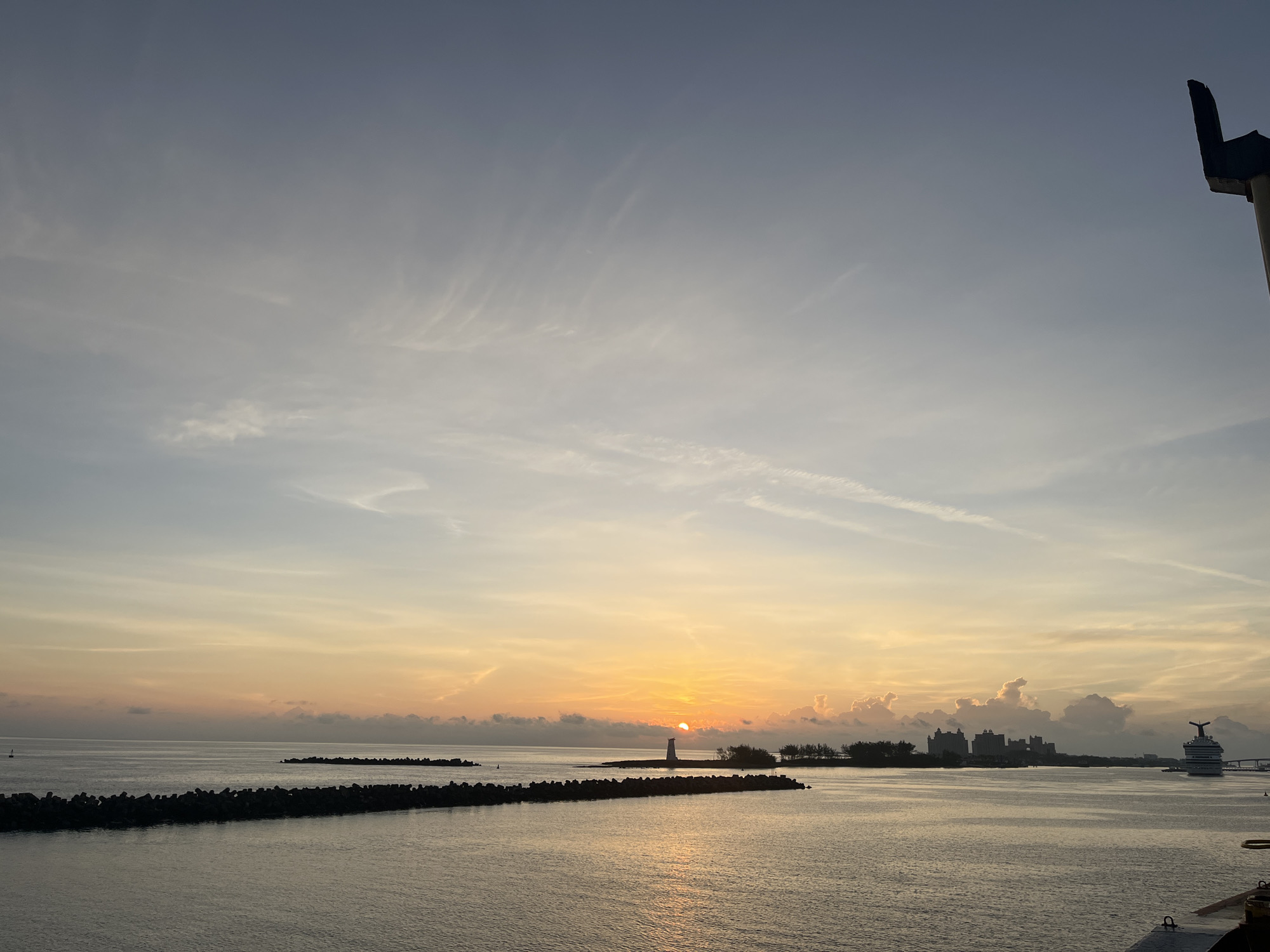 The image shows a sunrise or sunset over the ocean. A breakwater made of large rocks runs horizontally across the lower portion of the frame. A small island is visible in the center of the image, featuring a lighthouse. In the background, there is a cluster of buildings and a large cruise ship. The sky is filled with clouds, colored with hues of yellow and orange from the sun. The scene appears to be near a city, given the presence of large buildings, but the specific location is unidentified. The photograph seems to be taken from a high vantage point, possibly a ship or tall building.