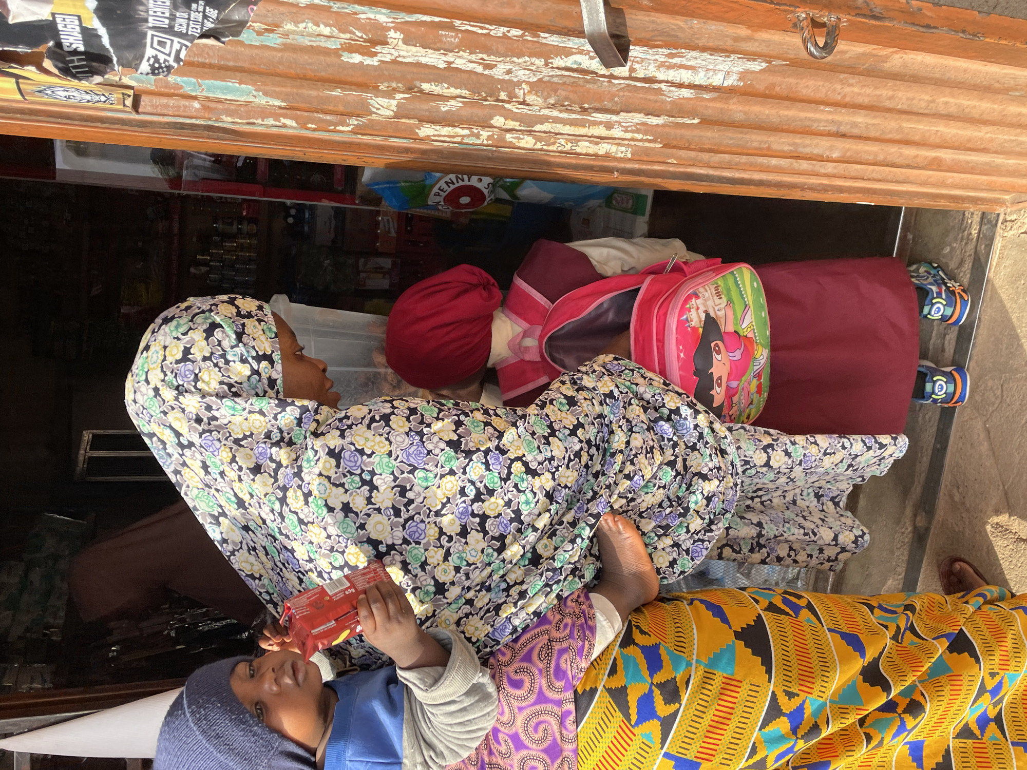 A woman wearing a floral headscarf and dress lies on a colorful fabric, presumably outdoors. A child, wearing a blue hat and shirt, lies beside her and holds a red package. Another child's red head covering and a pink Dora the Explorer backpack are also visible near the woman. The scene appears to be near a shop or storage area with a corrugated metal awning. Given the location and cultural clothing, the scene may be in Jos, Nigeria.