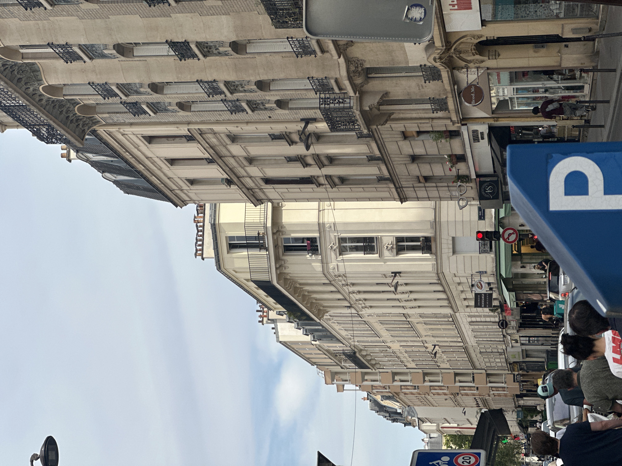 The image captures a street scene in Paris, France, likely during the daytime under an overcast sky. The perspective is from street level, looking down a cobblestone street lined with tall, multi-story buildings. The architecture is classic Parisian, with cream-colored facades, numerous windows with shutters, and ornate balconies with black iron railings. Some of the buildings have mansard roofs with dormer windows.
The street is moderately busy. Several cars are visible, including a silver hatchback and a black sedan. A few people are walking on the sidewalks, and some are seen in the doorways of shops. There is a blue parking sign with a white 'P' in the foreground, partially obstructing the view.
Various shops are identifiable on the ground floor of the buildings. One sign reads "MASSAGE," and another appears to be for a business called "Brood.Me." Other signs are partially obscured or difficult to read. Traffic lights are visible, indicating a street intersection. A street lamp is also present on the left side of the image. The overall impression is of a typical urban environment in Paris, with a blend of residential and commercial buildings.