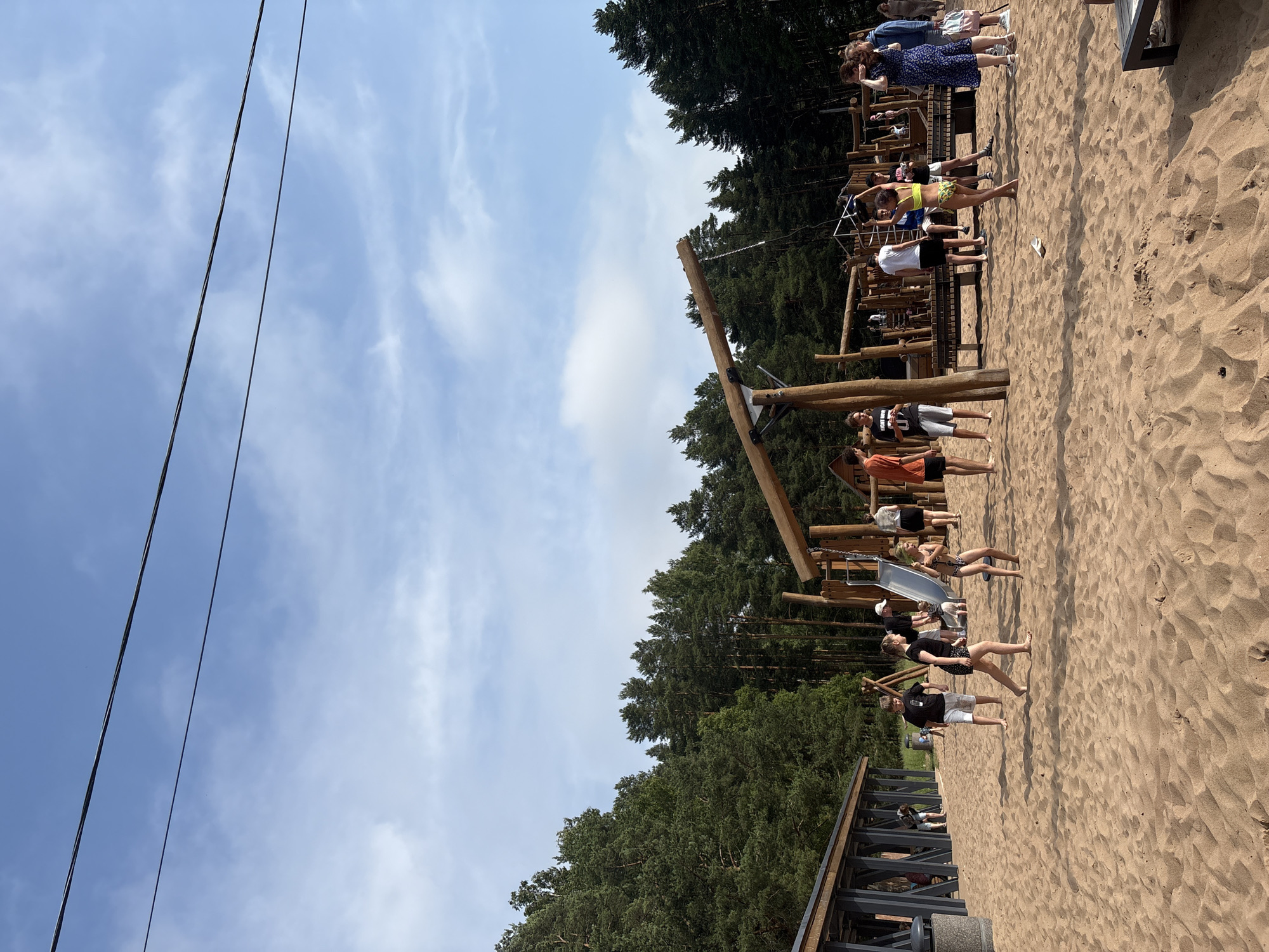 In Solnechnoye, Russia, on a sandy playground, children and adults are engaged in various activities. A playground structure made of wood dominates the center of the image, featuring a slide, swings, and climbing elements. Several children are playing on the playground equipment, while others are standing or walking on the sand. One girl in a yellow bikini is dancing, while a group of people are gathered near a wooden shelter on the left side of the image. A line of trees forms the backdrop, with a clear blue sky overhead. The sandy ground is littered with footprints and some small debris.