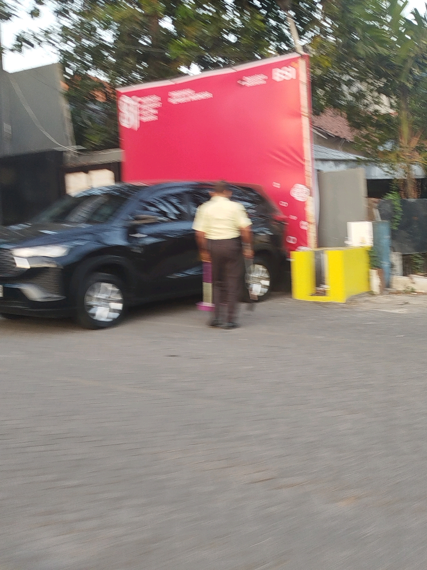 A person in a yellow shirt and dark pants stands on a paved area next to a black SUV. The SUV is parked in front of a large red banner with white text and graphics. To the right of the SUV, a bright yellow structure with a cutout is visible. In the background, trees and buildings can be seen. The scene appears to be outdoors, possibly during the day. The motion blur suggests the image was taken with a moving camera. The context provided indicates the location is Surabaya, Indonesia.