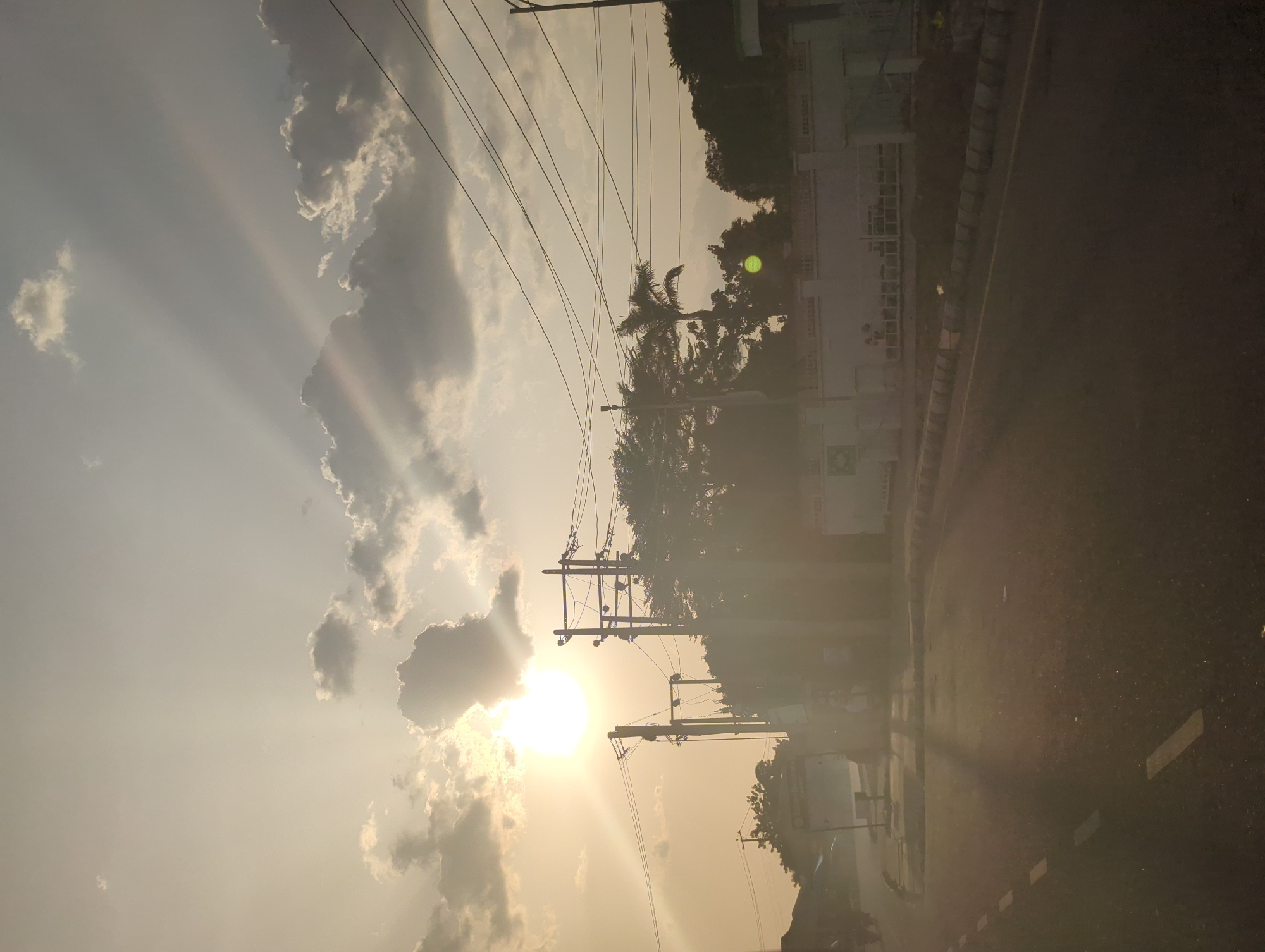 The photograph captures a vibrant sunset or sunrise scene over what appears to be a street or road in Barden Yana, Nigeria. The sky dominates the upper portion of the image, featuring a dramatic interplay of light yellow, orange, and grey hues with scattered clouds. The sun is a brilliant, intense orb, partially obscured by clouds in the lower-left area (when the image is viewed in landscape orientation), casting prominent crepuscular rays that fan across the sky, piercing through gaps in the clouds.
Several utility poles with a dense network of black power lines stretch horizontally across the mid-ground, silhouetted against the bright sky. Below these lines and to the right, numerous trees, including distinct palm trees and other dense foliage, are visible, mostly in silhouette, indicating a warm climate.
To the right side and extending towards the foreground, a dark asphalt road or street is visible, appearing underexposed due to the strong backlighting from the sun. It features white dashed and solid road markings, along with a grey curb or barrier running parallel to the road. Beyond the road, several buildings are partially visible, largely silhouetted against the brighter sky, with one structure exhibiting a faint green sign, though no text is discernible on it.
No people or specific activities are evident in the frame, allowing the natural light and urban landscape to be the primary focus. The strong backlighting creates a striking contrast between the luminous sky and the silhouetted elements of the environment. The overall impression is one of natural beauty and urban infrastructure bathed in the golden light of either dawn or dusk.