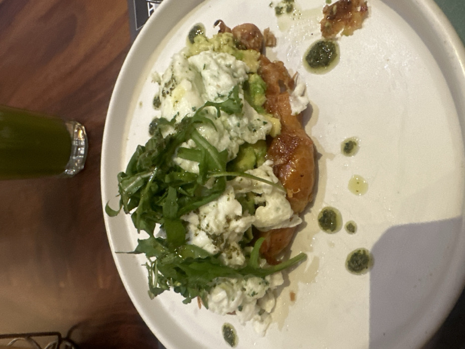 A white ceramic plate on a dark wooden table displays a breakfast or brunch dish. The central subject is an open-faced pastry, likely a croissant, appearing golden-brown and flaky. This pastry is topped with a layer of mashed or diced avocado, followed by a substantial amount of white, crumbled ingredient, resembling ricotta cheese or soft-scrambled eggs, speckled with green herbs. A generous pile of fresh, dark green arugula leaves crowns the dish. The plate's white surface features several small, distinct droplets of dark green pesto-like sauce and lighter yellow oil. In the upper background, out of focus, a clear glass contains a dark green beverage. The specific location within Mexico City cannot be determined from the provided image.