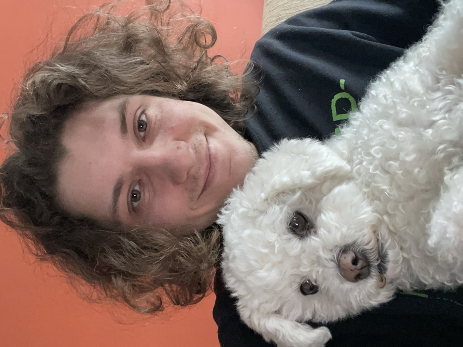 An indoor scene depicts a young adult male, characterized by light skin and curly brown hair, smiling and looking directly at the camera. He wears a dark sweatshirt. He is holding a small, white dog with curly fur, consistent with a Bichon Frise breed, which features dark eyes and a dark nose, and is oriented slightly to the viewer's left. The background consists of a plain orange wall on the left and a section of a light-colored, upholstered furniture item, possibly a sofa, on the right. The specific location within the city of Yekaterinburg, Russia, cannot be determined from the visual information provided.