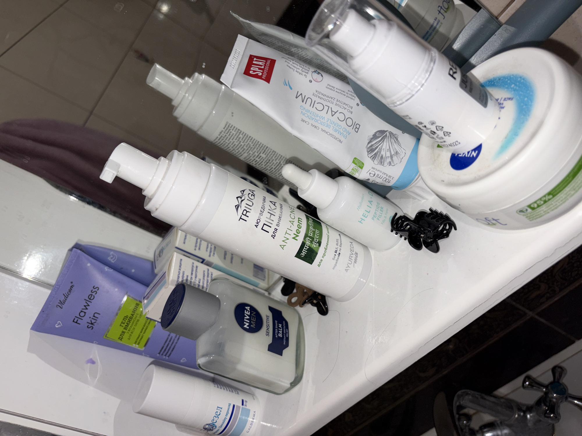 A top-down, medium shot captures a collection of toiletries arranged on a white surface, possibly a bathroom counter. The toiletries include various skincare and oral care products. A purple tube labeled "Vladicon Flawless skin" is prominent, alongside a clear bottle with a grey cap, identified as "NIVEA MEN SENSITIVE AFTER SHAVE BALM". Another white pump bottle, bearing the brand "TRIUGA", contains a product with Russian text and "ANTI-ACNE Neem". A tube of "SPLAT PROFESSIONAL BIOCALCIUM ENAMEL RESTORATION AND GENTLE WHITENING" toothpaste is also visible, as is a smaller white dropper bottle with "HELIA-D Peptide Filler". A large white tub with a blue and white label featuring "95%" is partially visible in the foreground. Two black hair clips are placed near the toiletries. The reflective surface suggests a mirror in the background, showing a blurred reflection of a purple towel. The overall environment appears to be indoors, likely a bathroom, with evidence of daily use.