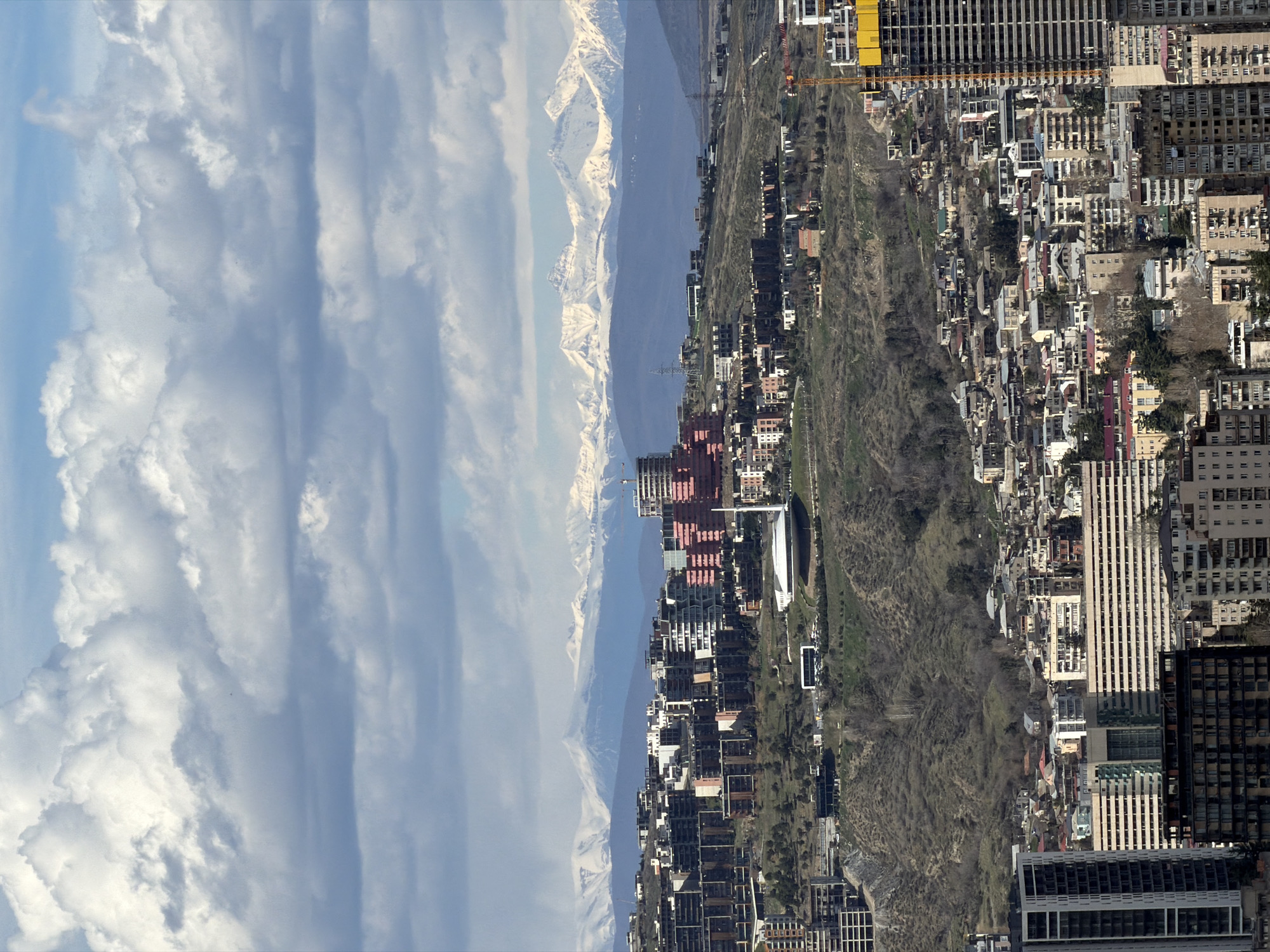 The image presents a cityscape with a dramatic view of Tbilisi, Georgia, oriented horizontally. The sky, filled with dynamic cloud formations, occupies the left side of the frame. Adjacent to the clouds are distant, snow-capped mountains, providing a natural backdrop to the urban setting.
Moving rightward, the cityscape unfolds, showcasing a mix of modern high-rise buildings and traditional architecture. A notable green belt, likely a park or forested area, bisects the built environment, offering a contrast between the natural and urban landscapes. The architecture consists of residential blocks, commercial towers, and individual houses nestled in the folds of the terrain. Construction cranes indicate ongoing development in certain areas. The image's composition emphasizes the interplay between the city's built environment and its natural surroundings.