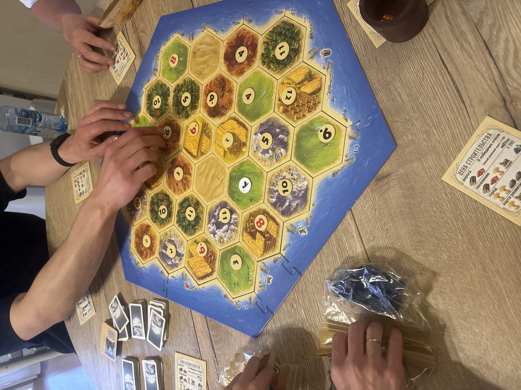 The image shows a game of Catan being played on a wooden table. Two people are visible, their hands interacting with the game components. The Catan game board is in the center of the image, with hexagonal tiles depicting different resources. A brown dice cup sits to the right of the board. Cards, some face up and some face down, are scattered around the board. A plastic bag containing game pieces sits near the bottom right corner. A water bottle is visible to the left of the board. A card displaying building costs is also present. The scene is well-lit, suggesting an indoor environment.