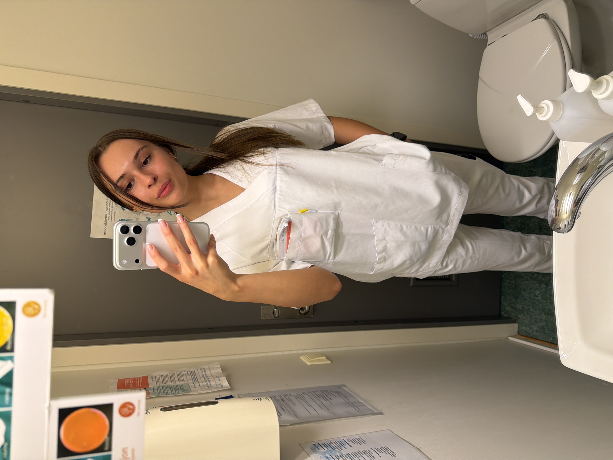 A young Caucasian woman with long brown hair tied in a ponytail is taking a mirror selfie in a bathroom. She is wearing white medical scrubs and appears to be in a healthcare setting. The bathroom is equipped with a toilet, a sink with a faucet, and several dispensers and papers on the wall. The flooring is a speckled green. The image is taken in a mirror, reflecting the woman and the surrounding environment. There is no visible indication of the specific location within Harstad, Norway.
