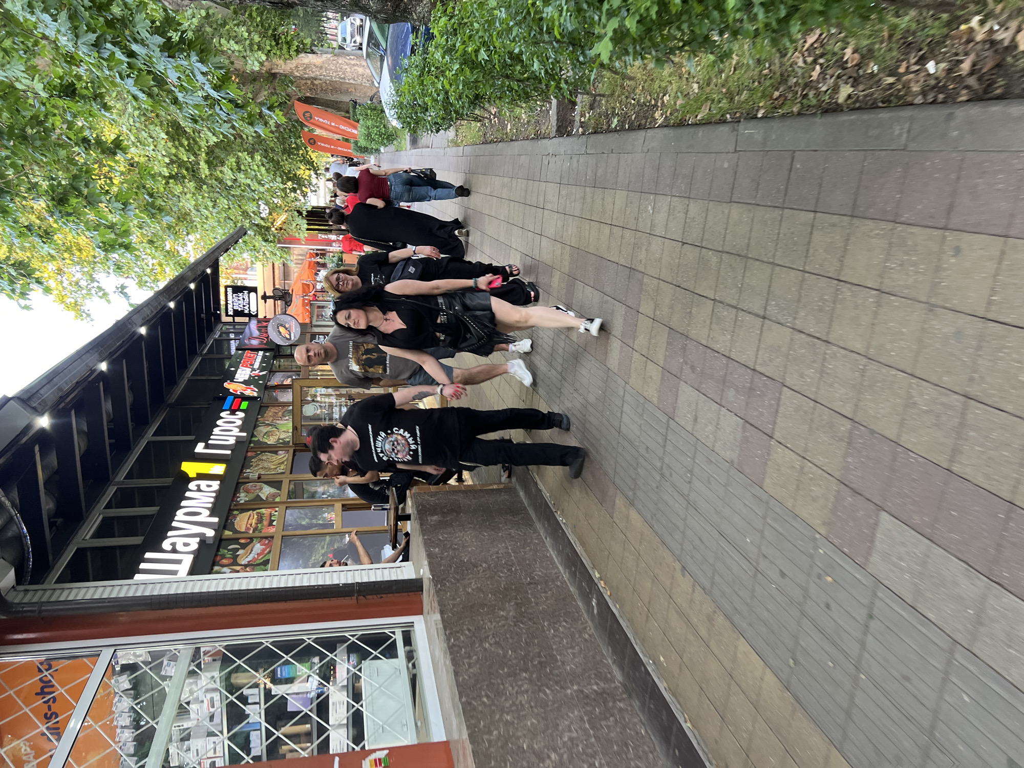 The image shows a street scene in Krasnodar, Russia, featuring a sidewalk lined with shops and trees. In the foreground, a group of people are walking on a paved sidewalk. The group includes several men and women, some dressed casually and others in darker, more alternative attire. A woman in a black leather skirt and top, with distinctive makeup, is prominent in the center of the group, walking towards the viewer. Behind her, others are walking in the same direction. To the left of the sidewalk is a building with a large glass window displaying various items and a sign with Cyrillic lettering that reads "Шаурма 1 Гирос" (Shawarma 1 Gyros), indicating a food establishment. The building has an awning extending over the sidewalk. To the right of the sidewalk, there are trees with green foliage and a grassy area, with a street and parked cars visible in the background. The overall scene depicts a casual urban environment.