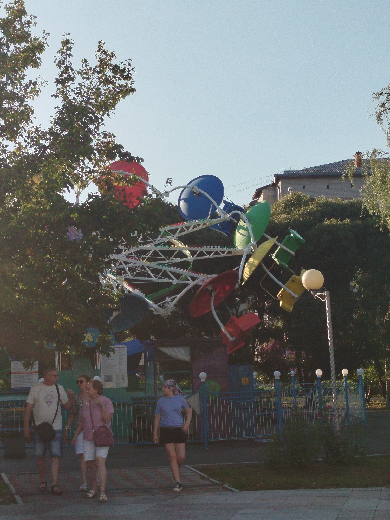 The image depicts an outdoor scene, likely an amusement park, with a vibrant Ferris wheel-like ride featuring colorful gondolas. The ride is partially obscured by lush green foliage from trees. In the foreground, a group of four people are walking along a paved path. A man in a light-colored t-shirt and denim shorts is on the left, followed by two women in casual attire. A young girl wearing a blue t-shirt and a headscarf walks ahead of them. Behind the people, a blue metal fence with decorative white finials encloses a pathway. Further in the background, a two-story brick building with a tiled roof is visible, suggesting a residential or administrative area. The sky is clear and bright blue, indicating daytime. The overall impression is a sunny day in a park. Given the context of the prompt, this scene is likely located in the city of Yalutorovsk, Russia.