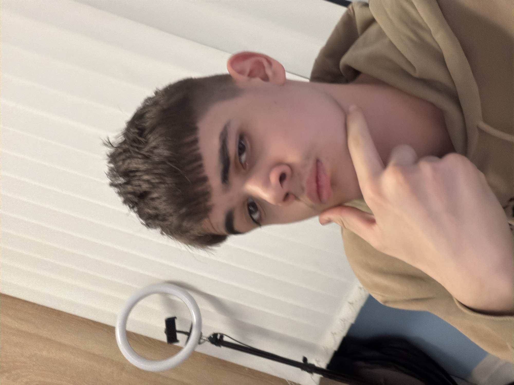A young man is looking at the camera. He has short, dark hair with bangs. He wears a tan hoodie. He is touching his face with his right index finger. A ring light is visible in the lower-left corner. A wall with white, horizontal slats is in the background. There is no evidence to suggest this image was taken in Pernik, Bulgaria.