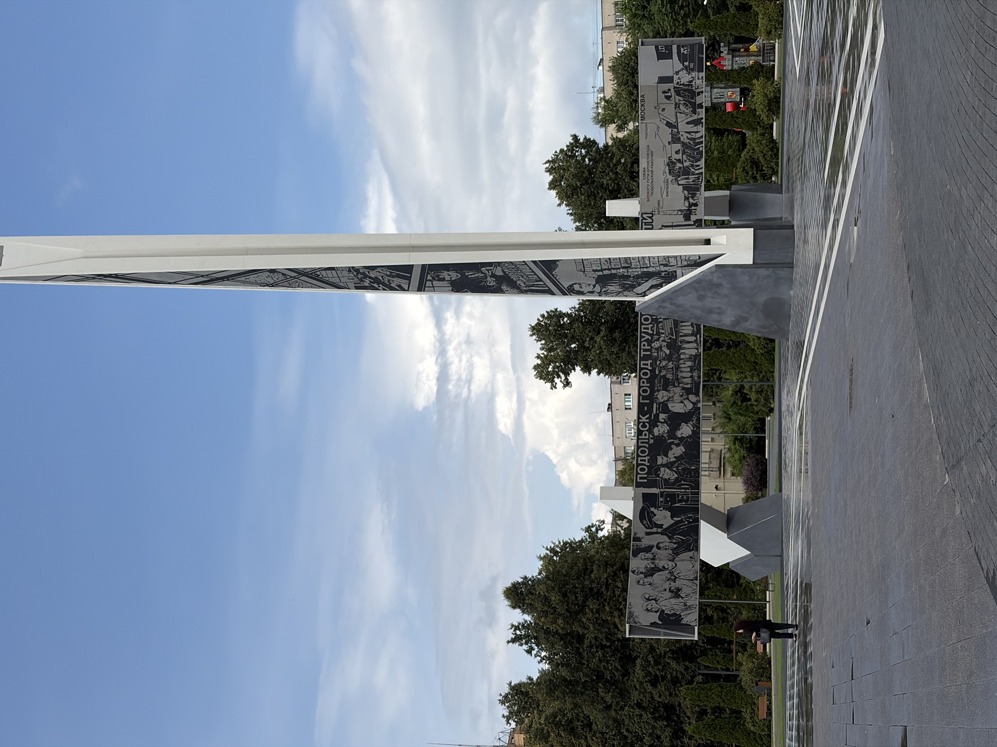 The image captures a monument in Podolsk, Russia, dedicated to the city's labor. The monument is a tall, white, angular structure adorned with black and white murals depicting scenes and people. The murals feature portraits, groups of individuals, and a map with text in Russian, indicating historical events or journeys related to the city.
In the foreground, a dark-paved public square is visible, wet from recent rain, with reflections on its surface. A solitary individual, dressed in dark clothing and carrying a bag, stands on the left side of the square, facing away from the monument.
Behind the monument, a line of dense green trees forms a backdrop. Beyond the trees, buildings and the sky are visible. The sky is partly cloudy with patches of blue. The overall impression is one of a somber yet proud memorial in an urban setting.