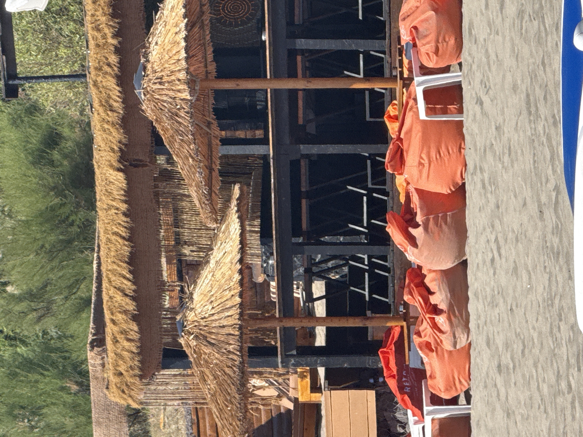 This is an outdoor photograph of a beachside establishment, likely a bar or restaurant, in Sozopol, Bulgaria. The scene features a sandy beach in the foreground with several large, orange beanbag chairs and white plastic stools arranged for seating. Behind the seating area, there is a wooden structure with a thatched roof, providing shade. A dark-colored bar or counter is visible beneath the thatched roof, with rows of chairs or loungers set up behind it. Two straw umbrellas are positioned on the beach, offering additional shade. The background is filled with lush green foliage. The lighting suggests it is a sunny day.