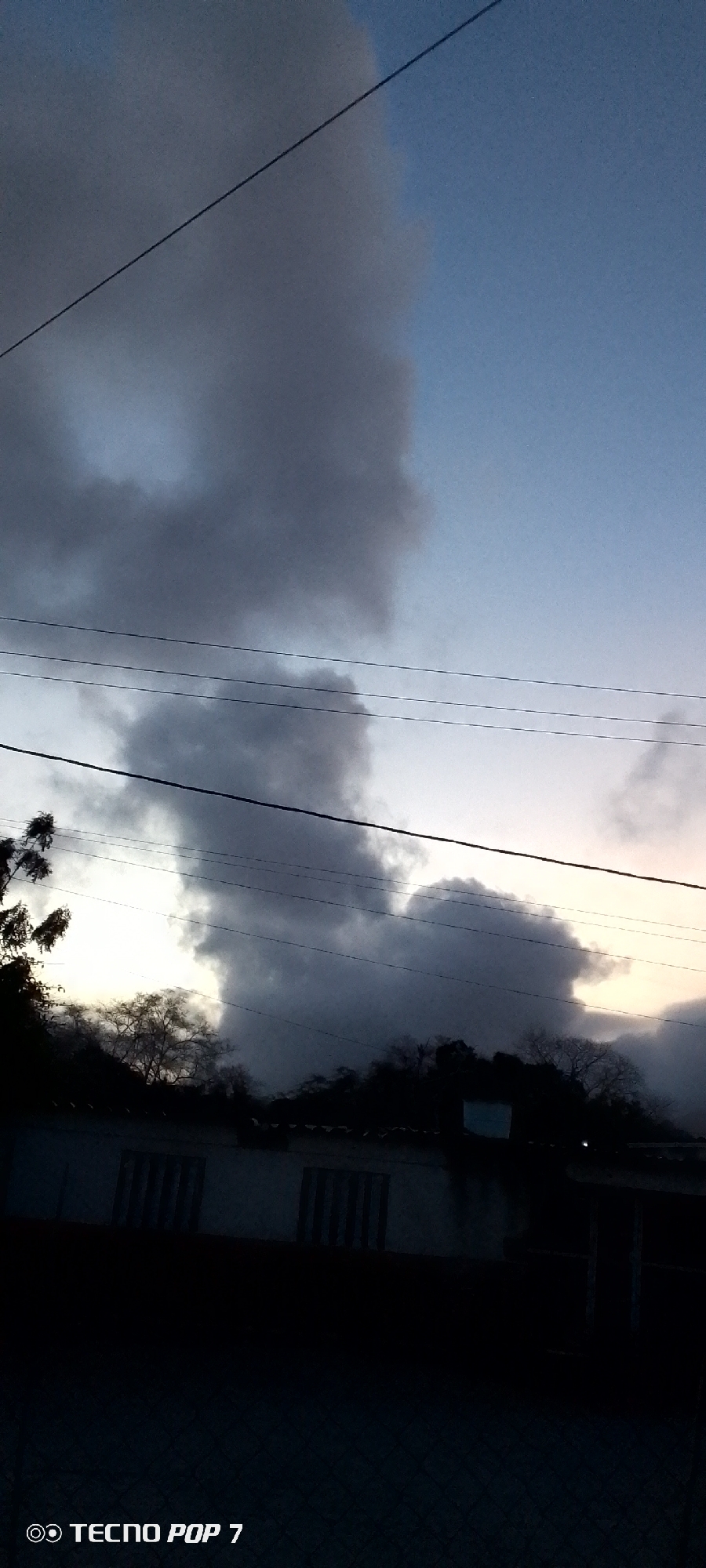 The image shows a dusky sky filled with heavy, grey clouds. The scene is partially obscured by the darkness of what appears to be a building roof and some trees silhouetted against the fading light. Power lines crisscross the upper portion of the frame. The bottom of the frame is mostly dark, suggesting it is either night or very late evening. The image was captured using a Tecno Pop 7 mobile phone. The location is in the city of Altagracia, Venezuela.