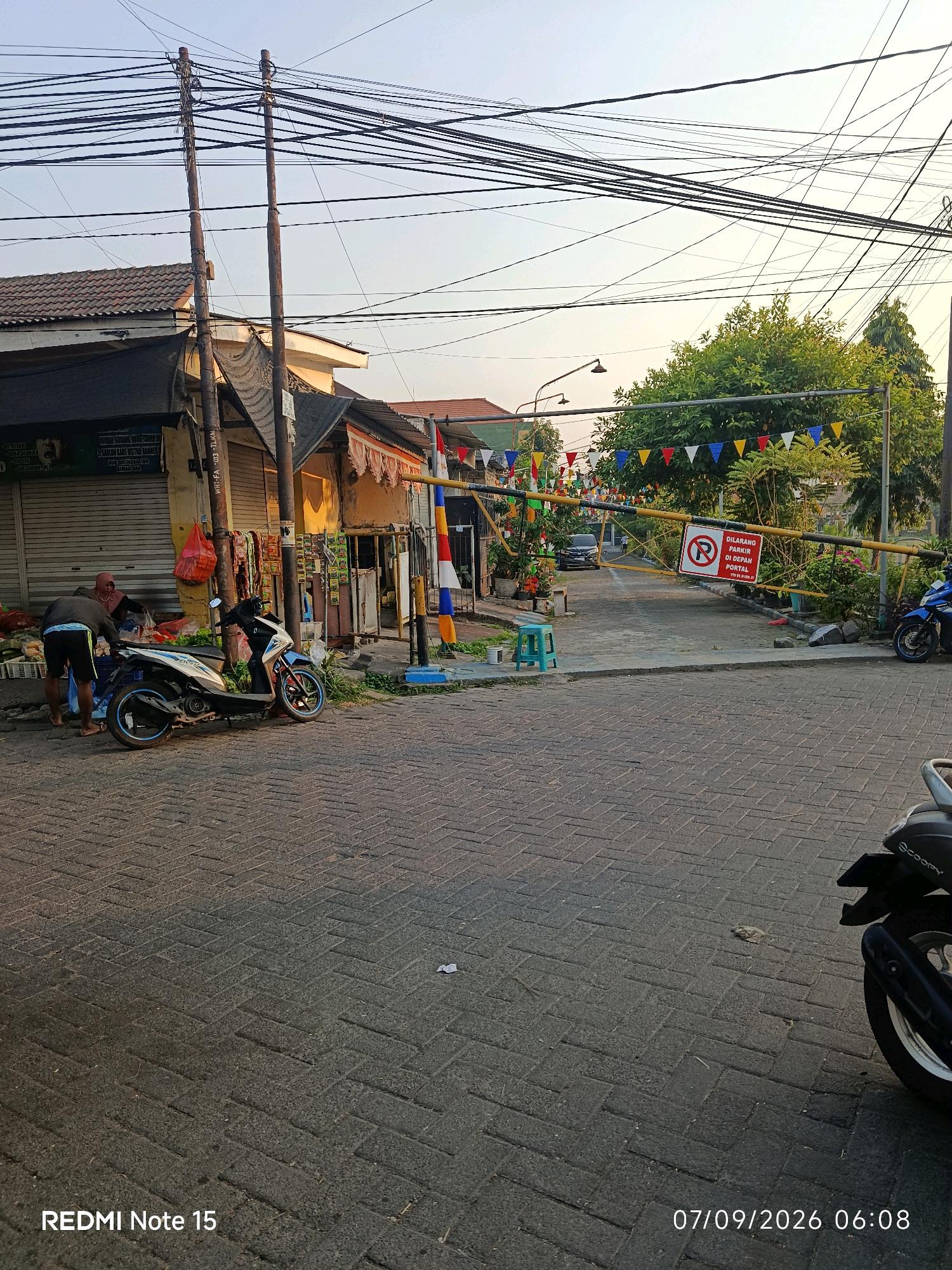 The image depicts a street scene in Keboansikep, Indonesia, at what appears to be dawn or dusk due to the warm, low-angle lighting. The foreground is paved with dark brick pavers arranged in a herringbone pattern. A black scooter is partially visible on the right side of the frame, with its rear wheel and exhaust pipe in focus. To the left, a white and blue scooter is parked.
Two individuals are present near the parked scooters. One, wearing a dark hoodie and black shorts with a blue waistband, is seen from behind. They appear to be interacting with a crate of goods or produce. Beside them, a woman wearing a pink head covering and a dark top is also engaged with the items. In front of them, a collection of fruits and vegetables is laid out, suggesting a market stall or roadside sale.
Behind the individuals and scooters, a row of small shops with rolled-down shutters and open fronts is visible. One shop has a yellow facade with some signage. Above the shops, a dense network of overhead electrical wires crisscrosses the sky, supported by two wooden utility poles.
Further down the street, a yellow barrier gate is lowered across the road, adorned with colorful triangular flags. A red and white circular sign with a red diagonal line through a symbol is attached to the gate. This sign indicates a prohibition, likely "No Parking" given the text below it which reads "DILARANG PARKIR DI DEPAN PORTAL." A dark-colored car is visible behind the gate, suggesting it is a traffic control point or entrance to a private area. Green trees and some potted plants are scattered along the roadside. The sky is a pale, hazy blue, with the sun not directly visible but its presence indicated by the soft light. The timestamp "07/09/2026 06:08" and "REDMI Note 15" are overlaid on the bottom of the image.