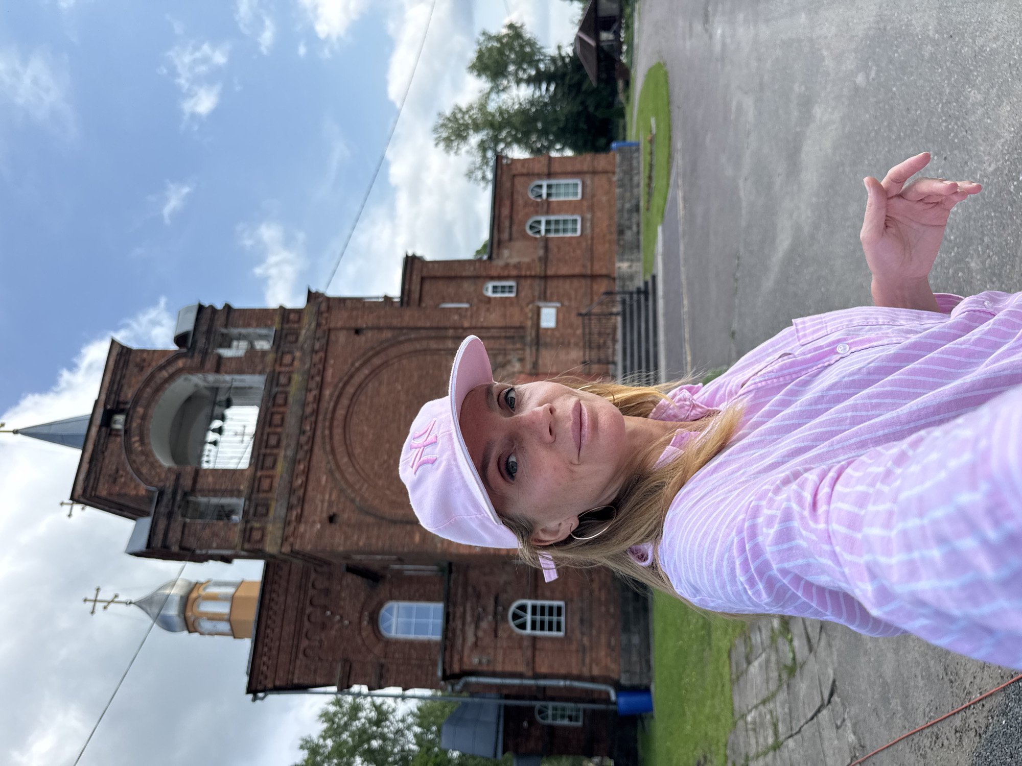 A blonde woman with freckles, wearing a pink and white striped shirt and a pink baseball cap with an embroidered logo, stands in front of a red brick church in Chudovo, Russia. She is looking towards the camera, with her right hand resting on her chest. The church has a bell tower with arched openings and a silver dome topped with a cross. The sky is cloudy with patches of blue. There is green grass and a paved area in front of the church.