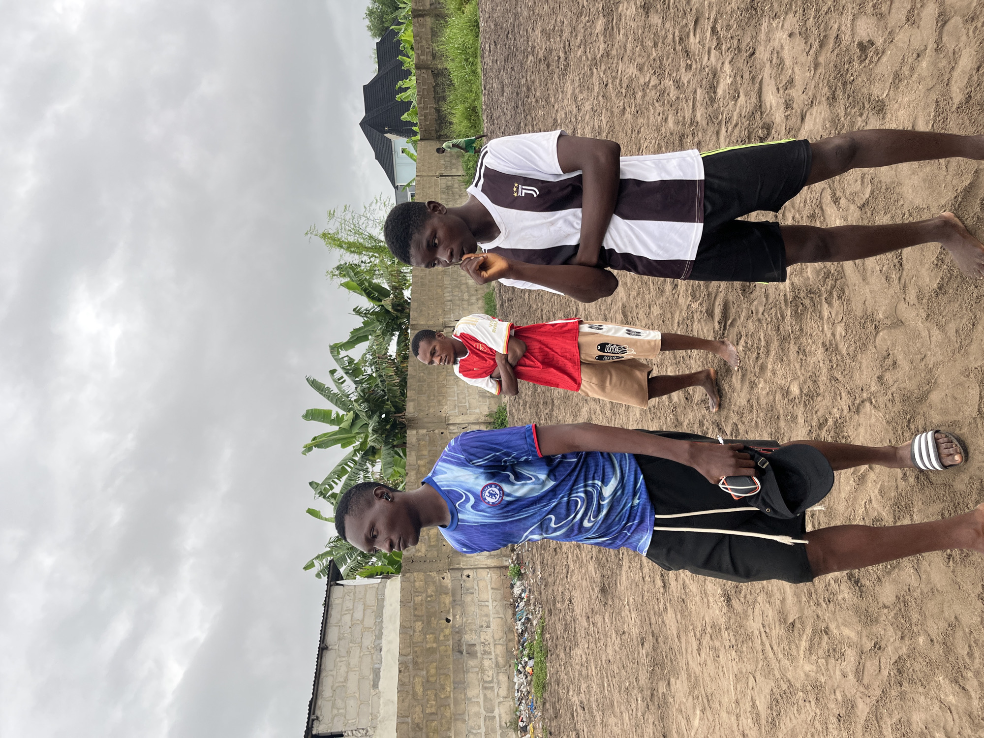 The image captures three young men standing on a sandy, uneven surface, likely a field or undeveloped area in Badagry, Nigeria. The sky above is overcast with dense, grey clouds. In the background, a low-lying, rough brick wall runs horizontally, with some debris visible near its base. Beyond the wall, lush green banana trees and other vegetation grow, suggesting a tropical climate. Further in the distance, a dark-tiled roof of a building is visible.
The man on the left is wearing a blue and white patterned t-shirt and black shorts. He is looking to his left with his left arm slightly bent. He is holding a black cap and a pair of sunglasses in his right hand. He is wearing white flip-flops with black stripes on his left foot, while his right foot is bare.
The man in the center is wearing a red t-shirt and tan shorts with a pattern. He is looking forward with his arms crossed. He is standing on the sandy ground with both feet bare.
The man on the right is wearing a black and white striped t-shirt and black shorts with a green stripe down the side. He is looking to his left with his right hand touching his chin and his left arm crossed over his chest. He is standing barefoot on the sandy ground. A fourth, much smaller figure, a young person in a green shirt and shorts, is visible in the far background, walking away from the camera. The ground is a mix of sand and packed earth, with some areas appearing damp.