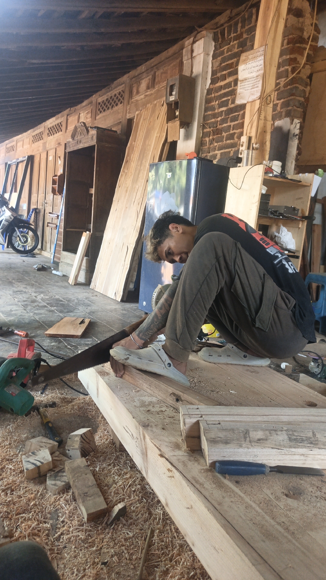 This image depicts a scene within a workshop, possibly in Kandangmas, Indonesia. The primary subject is a young man, with styled hair and tattoos on his arms, crouching down to saw a piece of lumber with a hand saw. He is wearing a dark t-shirt with a graphic design and olive-colored cargo pants. His feet are clad in light-colored clogs.
The workshop environment is cluttered with woodworking materials and tools. Large planks of wood are leaning against a wall, and sawdust is spread across the wooden floor. Various tools, including a green power saw and a chisel, are visible in the foreground. In the background, a refrigerator and shelves filled with items can be seen, along with brick walls and wooden ceiling beams. The lighting suggests it is daytime, possibly with sunlight filtering into the space. The overall impression is one of active construction or repair work.