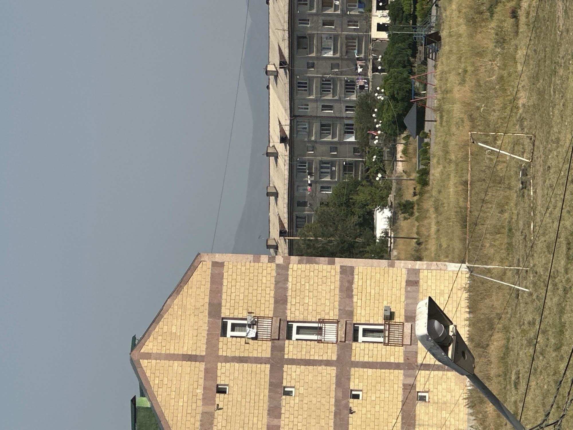 The image depicts an outdoor scene in Gyumri, Armenia. In the foreground, a street lamp extends diagonally across the frame, its shade angled upwards. Behind the lamp, a yellow brick building with maroon horizontal and vertical accents is visible. This building has multiple windows, some with small balconies, and laundry is hanging on one of the balconies. Further in the background, a long, grey, multi-story apartment building with a light-colored, sloped roof and numerous windows is prominent. Various trees and patches of dry grass are scattered throughout the scene. A small playground area with a swing set, slide, and a dark, umbrella-like structure is also visible. A rusty metal structure, possibly a goal for a sports field, is situated in the grassy area. In the far background, hazy mountains are discernible against a pale sky. The overall impression is one of an urban residential area on a clear day.