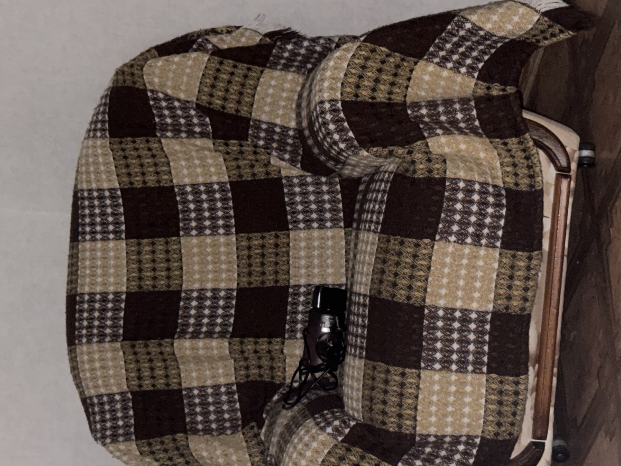A close-up shot captures a checkered armchair draped with a brown and beige throw blanket. A black digital camera with its cord lies on the seat of the chair. The chair is positioned on a wooden floor, with a hint of a wall visible in the background. There is no information in the image to suggest a location in Chelyabinsk, Russia.