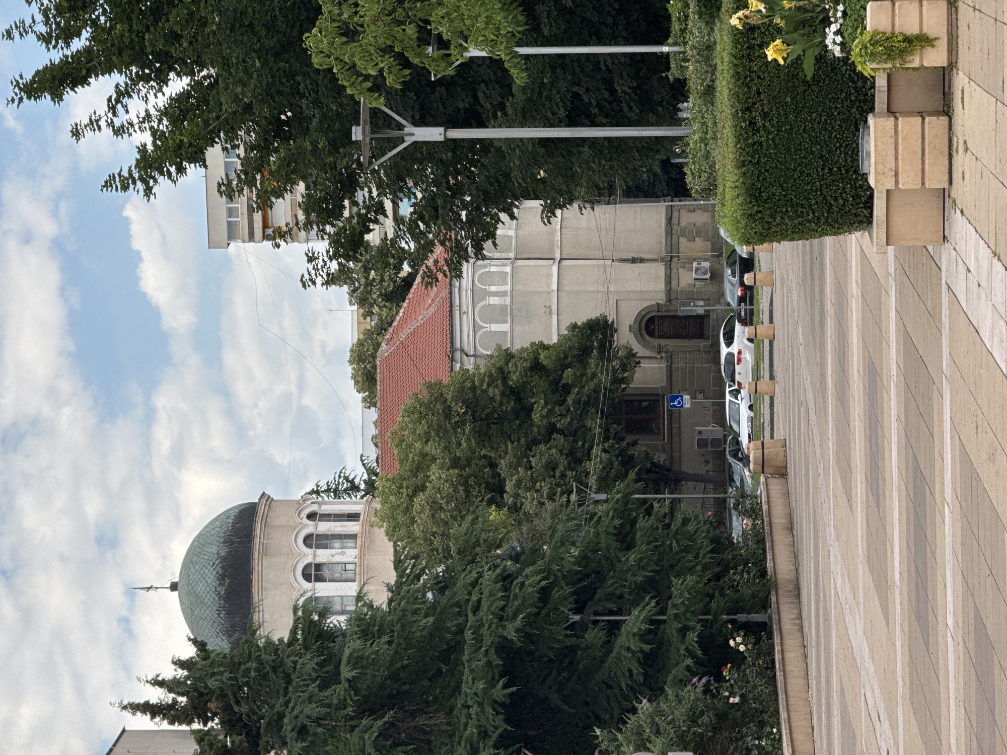 The image shows a daytime view of a public square with a building featuring a green dome and arched windows, likely a religious or historical structure, situated behind tall evergreen trees. Adjacent to it, another building with a red-tiled roof and arched facades is visible. In the foreground, a paved area with a geometric pattern of beige and white tiles leads towards the buildings. There are several parked cars, street lamps, and carefully manicured hedges and planters with flowers. A wheelchair accessibility symbol is visible on a sign near the second building. The sky is partly cloudy. This scene is identifiable as the area around the St. George Church in Yambol, Bulgaria, specifically the plaza in front of it.