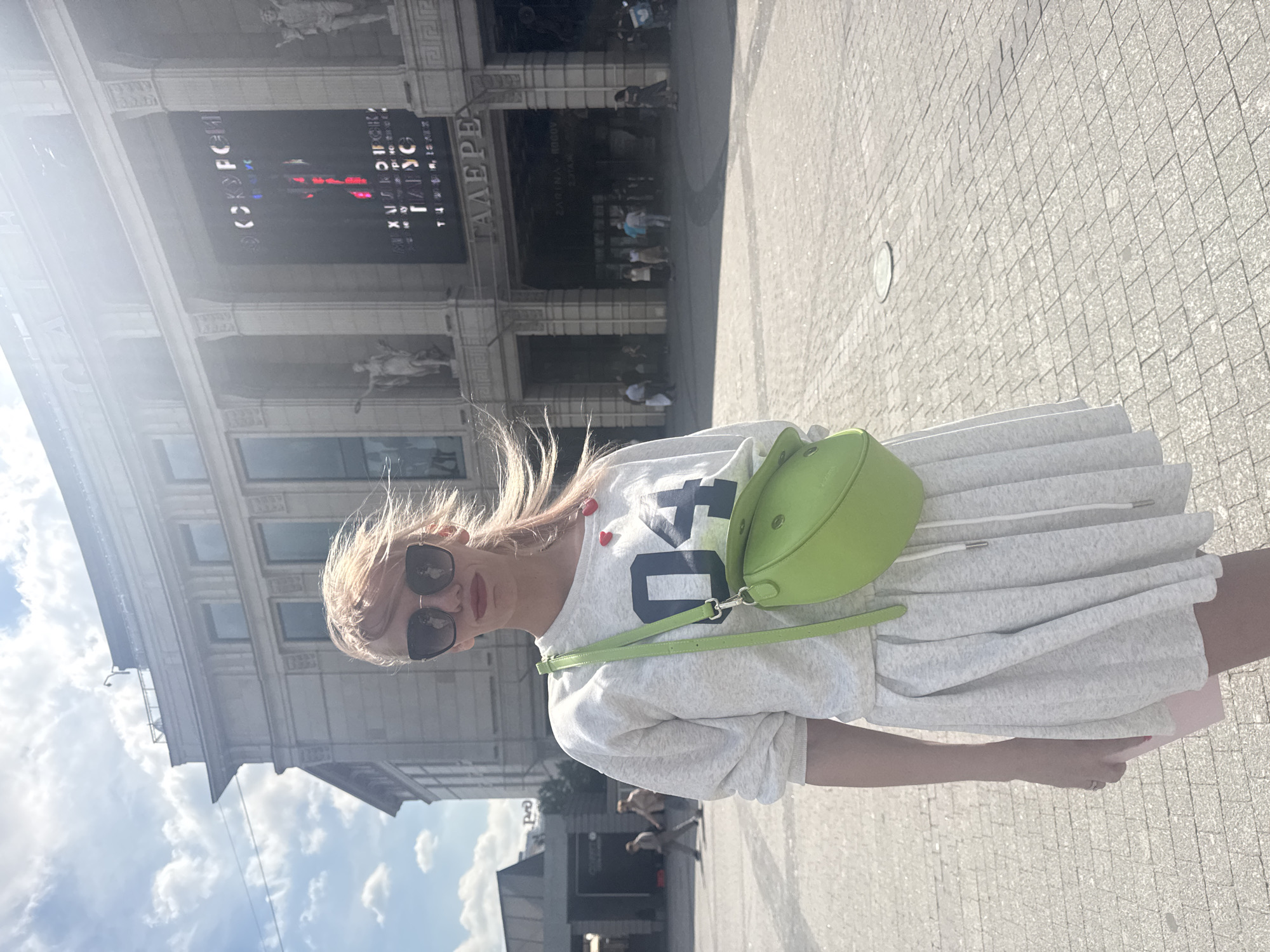 A young woman with blonde hair, sunglasses, and a light grey outfit stands in front of a large building with classical architecture, likely in Saint Petersburg, Russia. She is wearing a sweatshirt with the number "04" printed on it and a light grey pleated skirt. A bright green crossbody bag is slung over her shoulder. The building behind her has an illuminated sign that appears to advertise an event, with text that includes Cyrillic letters. Several people are visible in the background, walking in front of the building's entrance, some entering or exiting. The ground is paved with square stones. The sky is bright with scattered clouds, and the sun is casting strong shadows. The woman's hair is being blown by the wind.