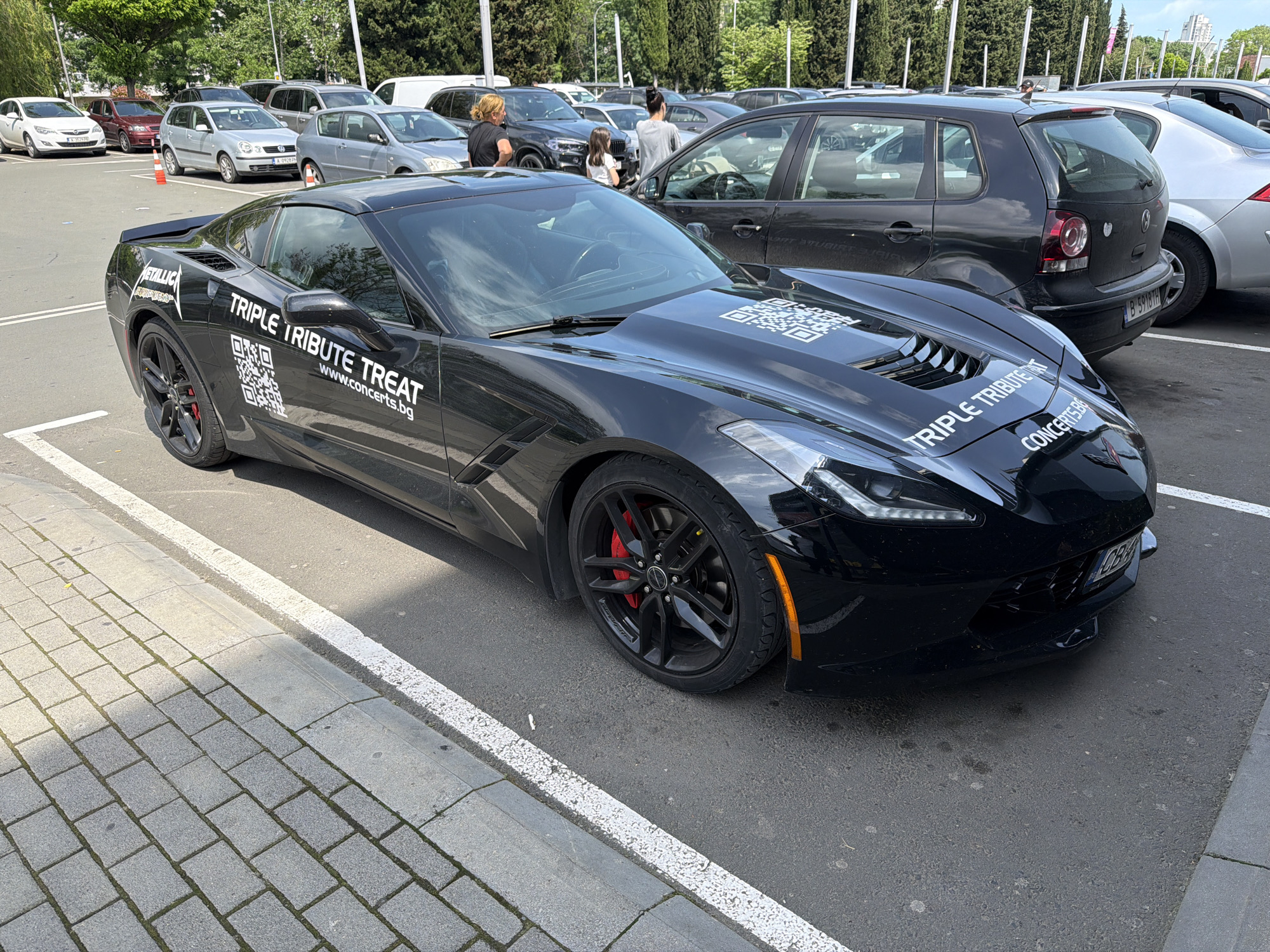 The image shows a black Chevrolet Corvette parked in a parking lot. The car has "TRIPLE TRIBUTE TREAT" and the website "www.concerts.bg" written on it, as well as a QR code. It also has a "Metallica" logo on the rear quarter panel. The Corvette has a Bulgarian license plate that reads "CB 6898 BH". Other vehicles are parked nearby, including a black Volkswagen Polo with the license plate "B 5918TH". In the background, several people are seen walking. The pavement is made of rectangular gray stones. The image appears to have been taken in Burgas, Bulgaria, indicated by the Bulgarian license plates on the vehicles.