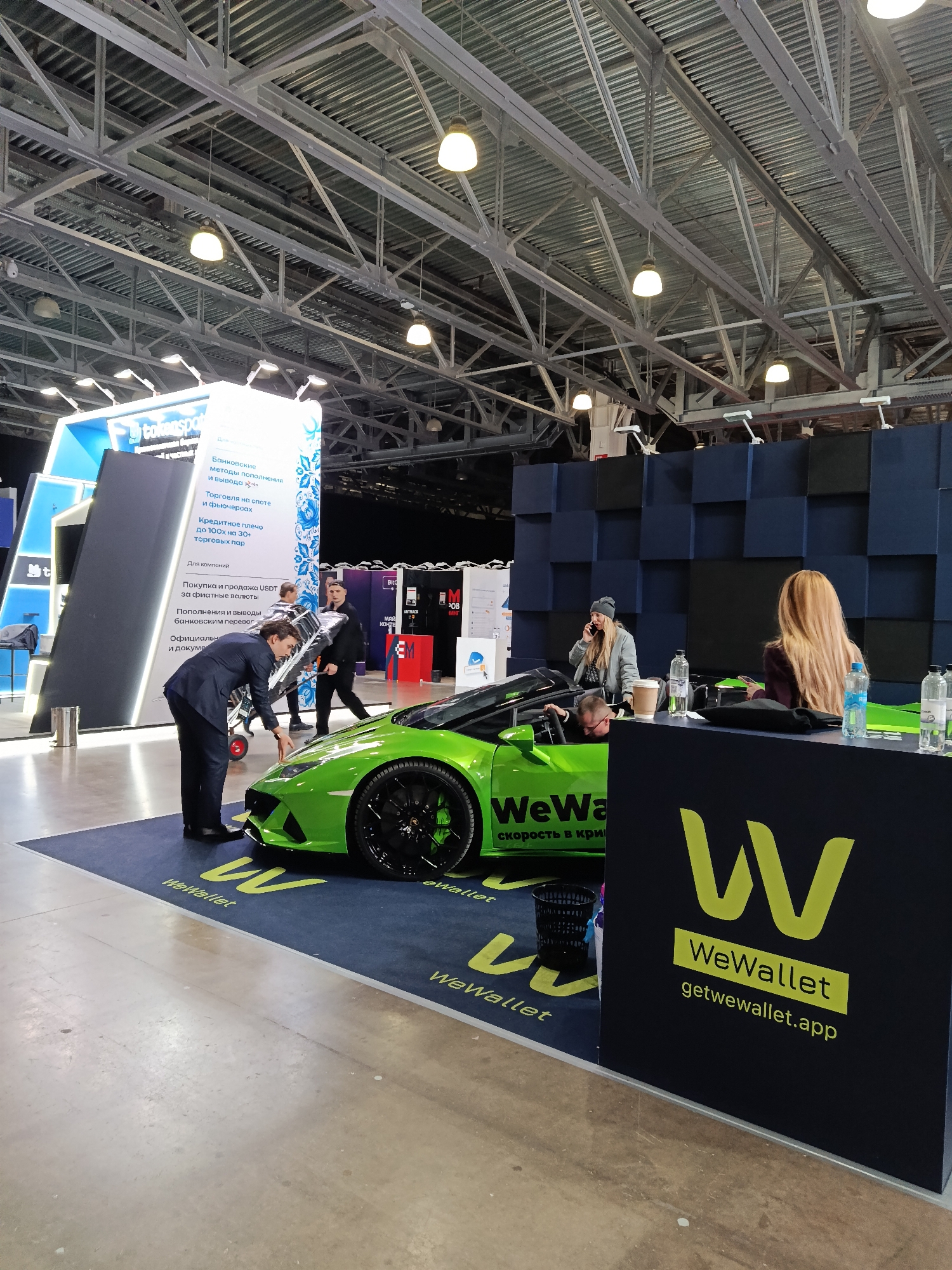 Here is a forensic description of the image:
The image shows an indoor exhibition space. A bright green Lamborghini convertible is on display on a dark blue carpet bearing the "WeWallet" logo. A man in a suit is standing near the front of the car, appearing to be adjusting something. Behind the car, a woman in a beanie is talking on the phone. Another woman with long blonde hair is seated at a desk with water bottles on it. The desk has the "WeWallet" logo and website address displayed. A black wastebasket is next to the car.
In the background, there are various exhibition stands. One stand has text in Russian with the words "Банковские методы пополнения и вывода" (Banking methods of replenishment and withdrawal). The ceiling is high with exposed metal beams and lighting fixtures.