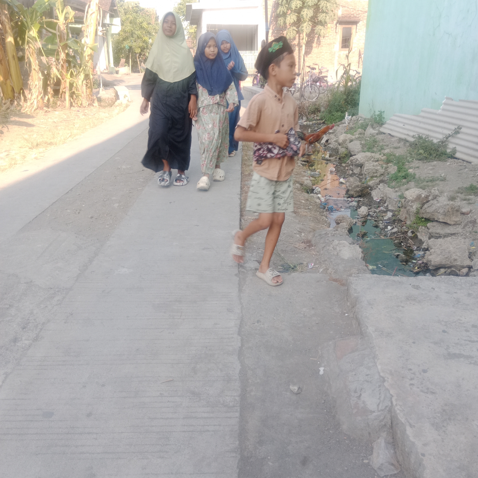 In the city of Sidorejo, Indonesia, a group of four children walk along a concrete pathway. The first child on the left is wearing a light green hijab and a long black dress. Next to her is another child in a dark blue hijab and a floral patterned dress. Following them is a child in a blue hijab. The child closest to the foreground is a boy wearing a light brown collared shirt and patterned shorts, with a dark green cap. He is walking with his head turned to the right and appears to be holding a piece of cloth. To the right of the pathway, there is a small, dirty ditch filled with water and debris, including plastic waste. Beyond the ditch, rocks and vegetation are visible. In the background, a light blue wall and a brick building with a window can be seen. There are also bicycles parked in the background, suggesting a residential or semi-urban area. The scene is illuminated by natural sunlight.