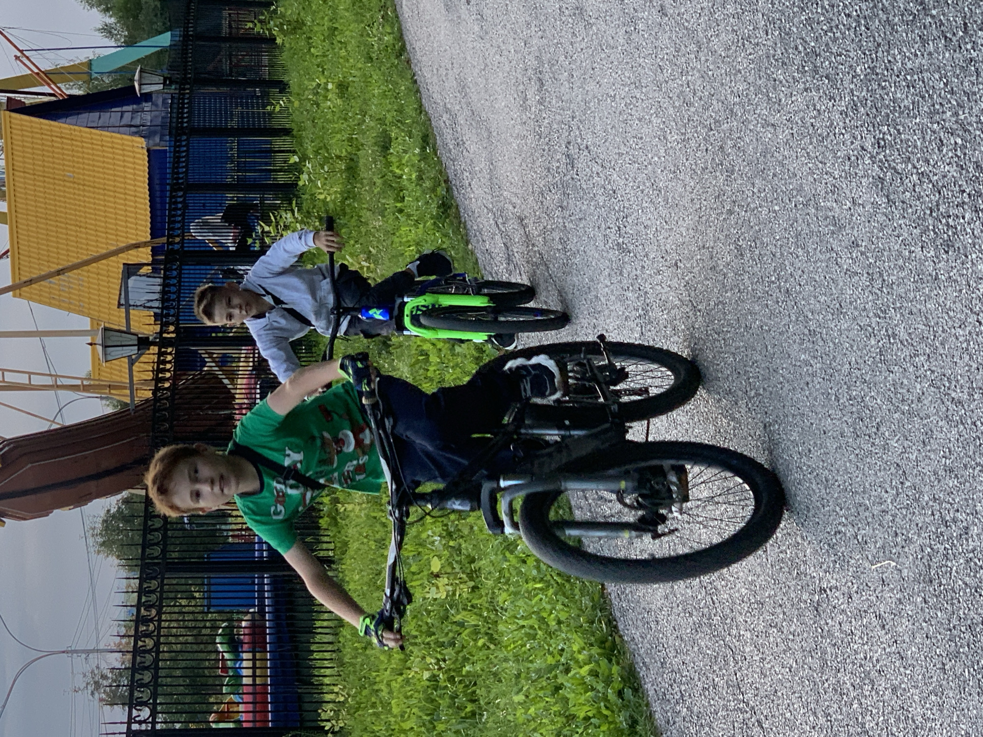 The image shows two young boys on bicycles riding on a paved path. The boy in the foreground is wearing a green t-shirt and dark pants, with a black strap across his chest. He is looking forward and appears to be in motion. The boy behind him is wearing a light gray hooded sweatshirt and dark pants, also appearing to be riding his bike. They are in an outdoor environment with green grass and trees visible. In the background, there is a black fence and some structures that appear to be part of a park or amusement area, including a building with a yellow roof and a tall, curved wooden structure. The lighting suggests it is daytime, possibly late afternoon or early evening. The location is identified as Prokopyevsk, Russia.