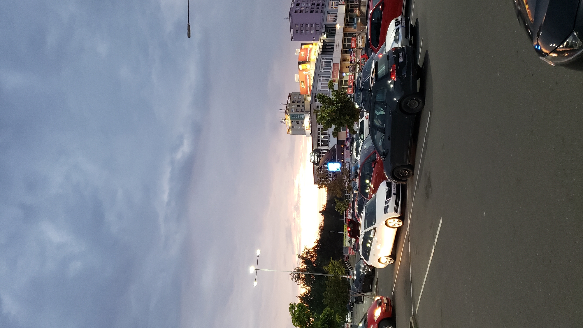 The image depicts a parking lot at dusk in Prešov, Slovakia. The sky is overcast with dramatic clouds tinged by the setting sun, casting a warm glow on the horizon. In the foreground, several cars are parked, including a white Skoda wagon illuminated by its own headlights. Other vehicles visible are a red car in the left foreground, a dark gray Volkswagen wagon, a red hatchback, and a white car. In the background, a modern shopping center with visible signage such as "RANC ADMIRAL" and "SUPERMARKET" stands. Trees line the edge of the parking lot, and several apartment buildings are visible further in the distance. A lamppost with multiple lights stands prominently in the middle ground, illuminating the parking area. The scene captures a typical evening in an urban environment with a mix of commercial and residential structures.