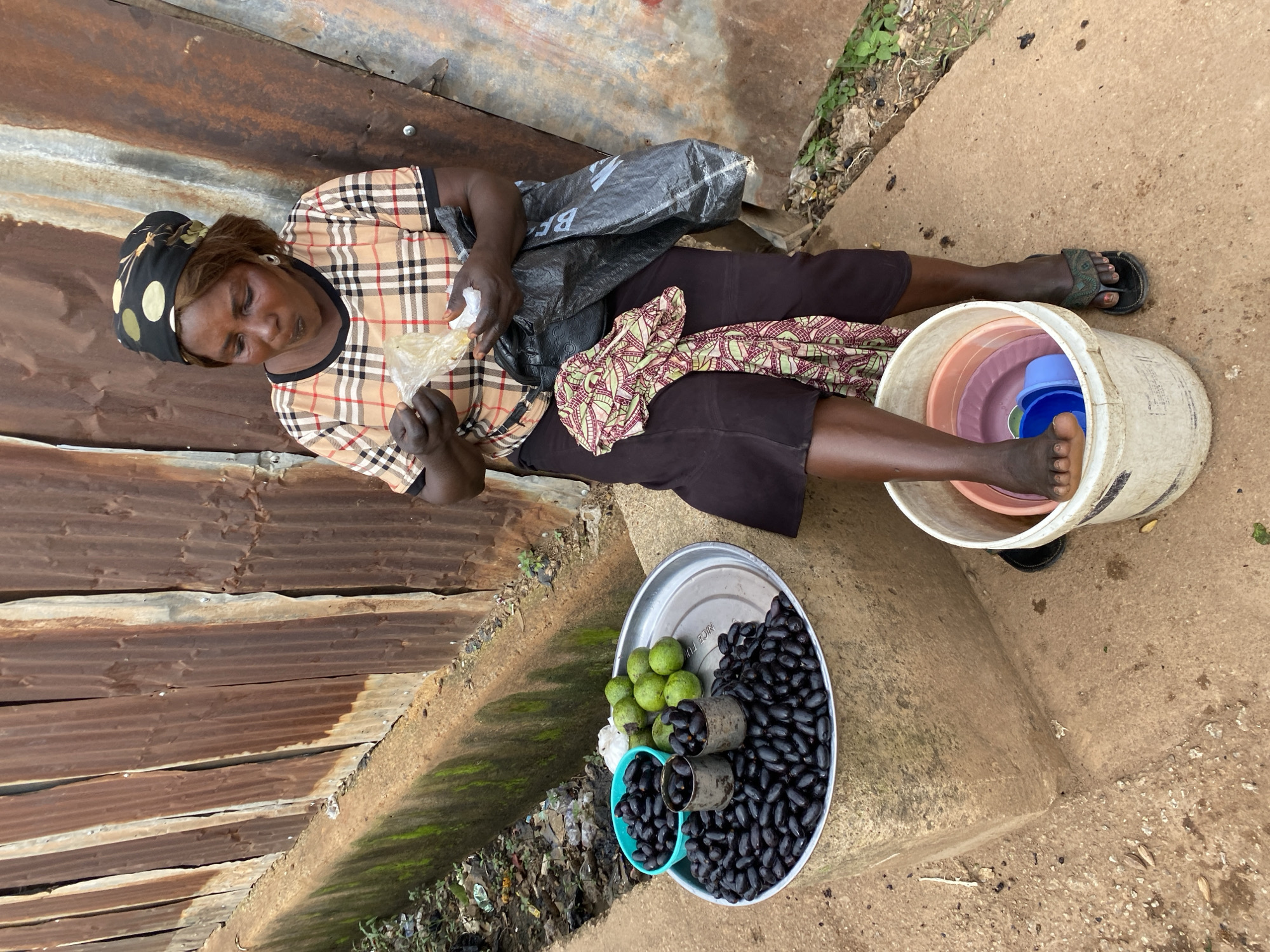 In Bukuru, Nigeria, a woman sits by a rusty corrugated metal wall, her gaze directed downwards as she prepares to eat from a small plastic bag. She is wearing a checkered shirt and a headscarf. In front of her, a metal tray holds a pile of dark, oval-shaped fruits, alongside a teal bowl filled with more of the same fruit and a smaller teal bowl containing green, round fruits. A white bucket sits to her right, containing stacked colorful plastic bowls. The ground around her is dirt and concrete.