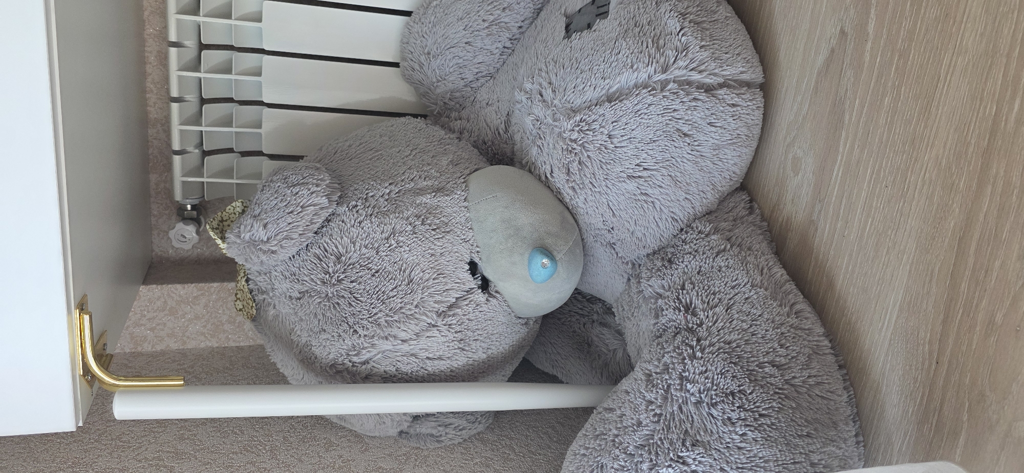 A large, plush, grey teddy bear is positioned in the lower right quadrant of the frame, resting on a light-colored wooden floor. The bear is facing slightly towards the left, with its head bowed and one arm resting in its lap. A small, blue, gem-like nose adorns its snout, and a decorative bow is visible on its head. To the left of the bear, a tall, white cylindrical object, possibly a pole or support, stands vertically. In the background, a white radiator with vertical slats is partially visible. Above the radiator and bear, a white shelf is supported by a gold-colored L-bracket attached to the wall. The wall behind the bear has a textured, light-brown wallpaper. The scene appears to be indoors, likely in a residential setting. The image does not provide enough information to determine a specific location within Sumgayit, Azerbaijan.