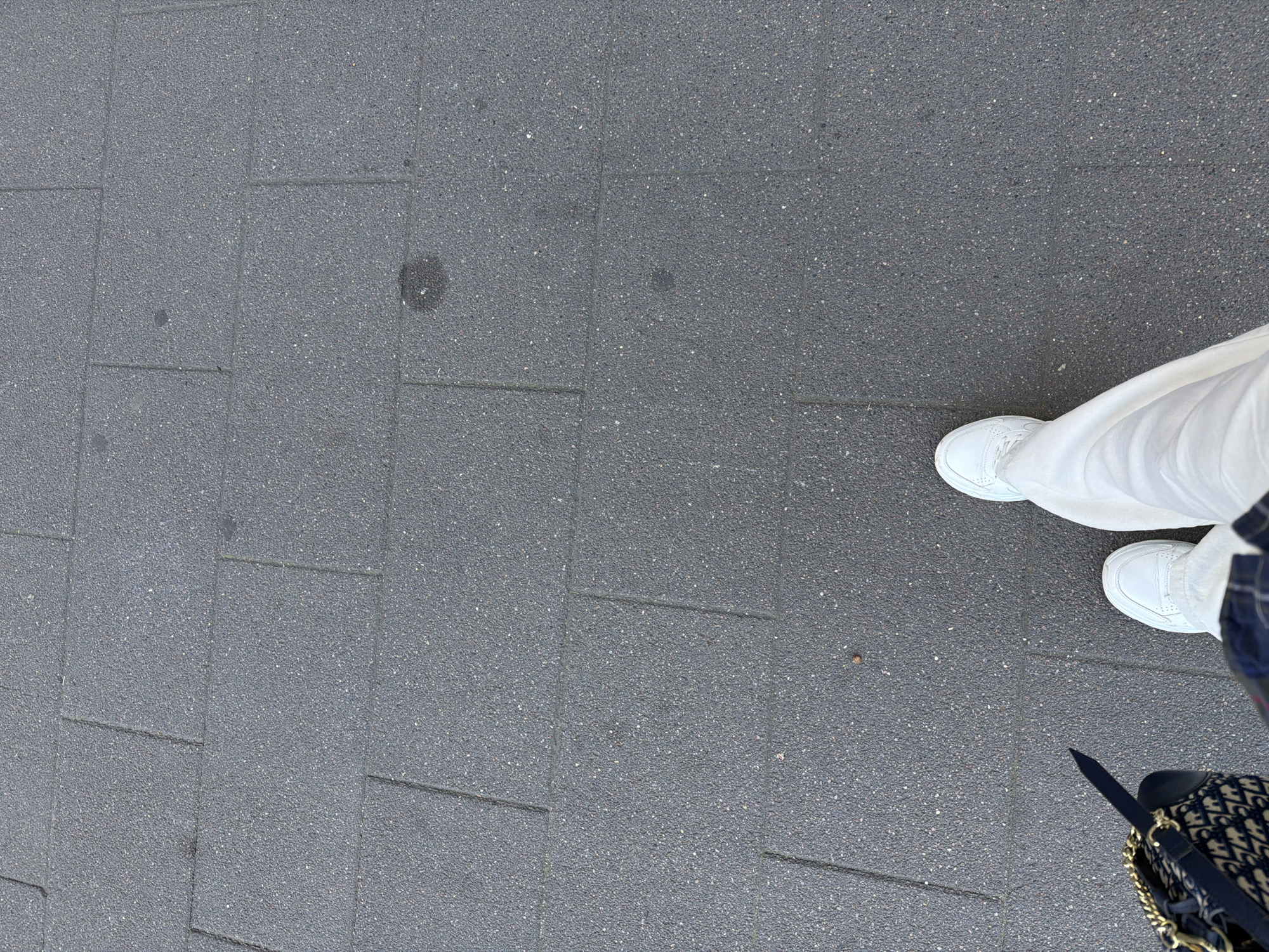 The image is a high-angle shot of a person's legs and feet walking on a sidewalk in Khimki, Russia. The sidewalk is paved with dark gray rectangular tiles. The person is wearing white sneakers and white pants. To the left, a portion of a navy blue and beige patterned bag with a gold chain strap is visible. The person's action is walking forward. The environment is an outdoor urban setting.