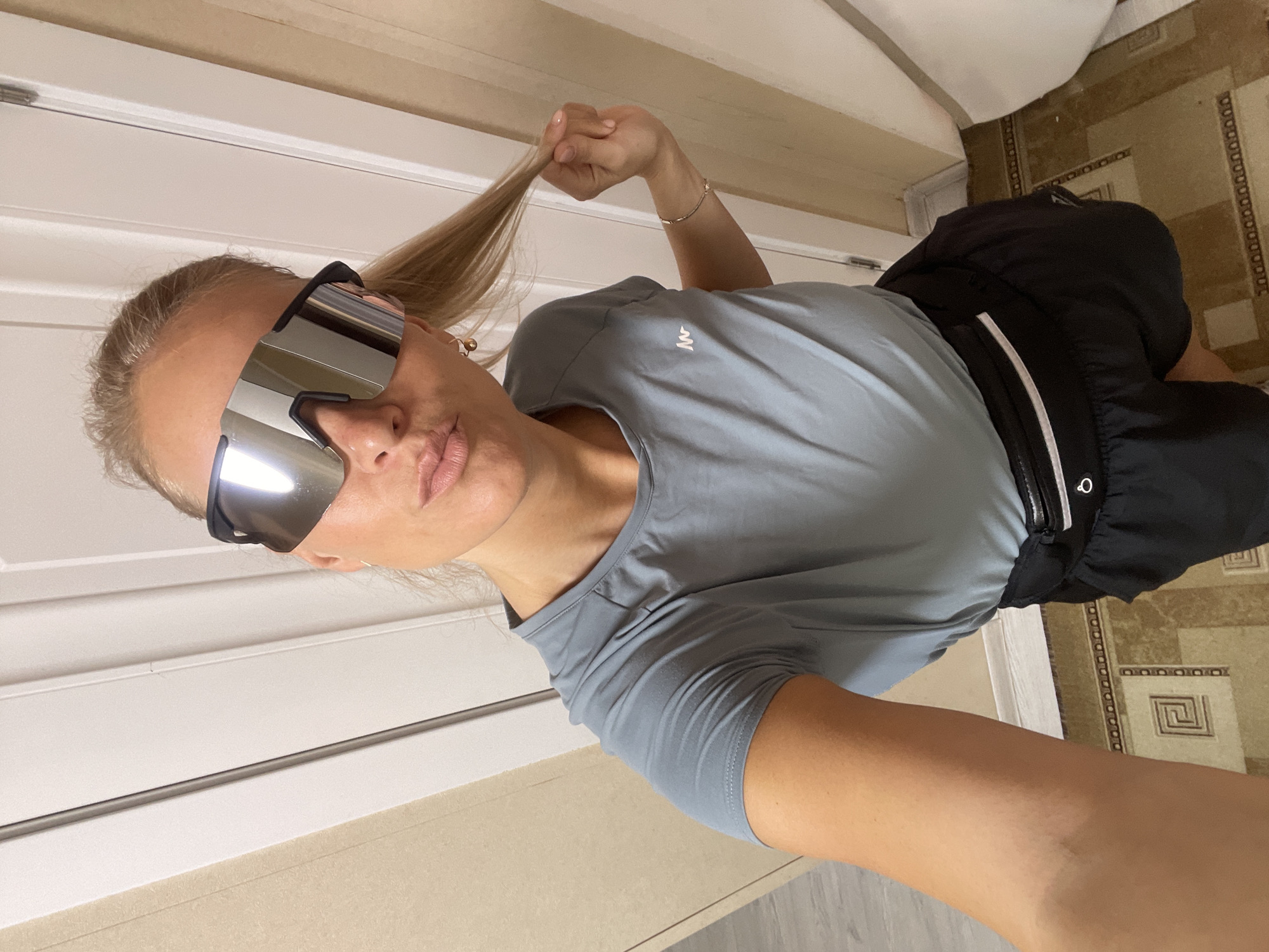 A woman is posing indoors for a selfie. She is wearing large, mirrored sunglasses, a gray t-shirt, and black shorts with a black fanny pack. She is holding a strand of her blonde hair. The background includes a white door, a section of wall, and a patterned floor. There's no indication of a location in the city of Baranovka, Russia.