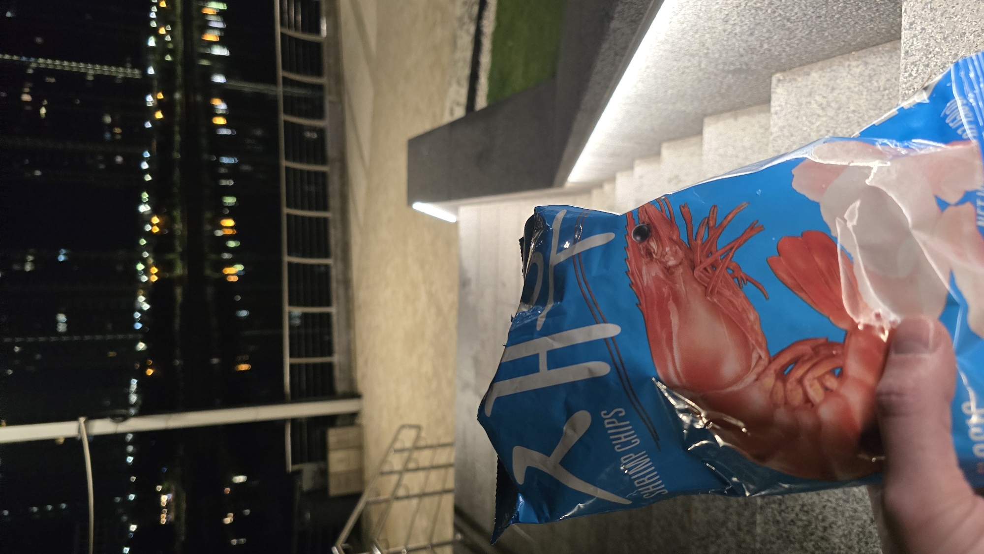 A person holds an open blue bag of "SHRIMP CHIPS" in the foreground, with a large image of a cooked red shrimp prominently displayed. Korean characters, likely "새우깡" (Saewookkang, a Korean shrimp snack brand), are visible at the top left of the bag. The bag is held by a thumb and fingers, obscuring the bottom portion.
The scene is set at night on the Dnieper River embankment on the right bank of Kyiv, Ukraine, looking towards the Left Bank's residential districts. In the midground, concrete steps with integrated horizontal strip lighting descend towards a paved area. A modern, dark-colored railing with an illuminated top edge runs alongside the stairs on the right. Another set of metal railings and stairs is partially visible on the left. Beyond the paved area, a dark body of water, the Dnieper River, reflects the blurred lights of distant city buildings. A metal fence or barrier separates the paved area from the river. In the background, numerous high-rise buildings on the opposite bank are illuminated, their lights appearing as bokeh. A strip of green grass is visible to the far right next to the illuminated railing.
